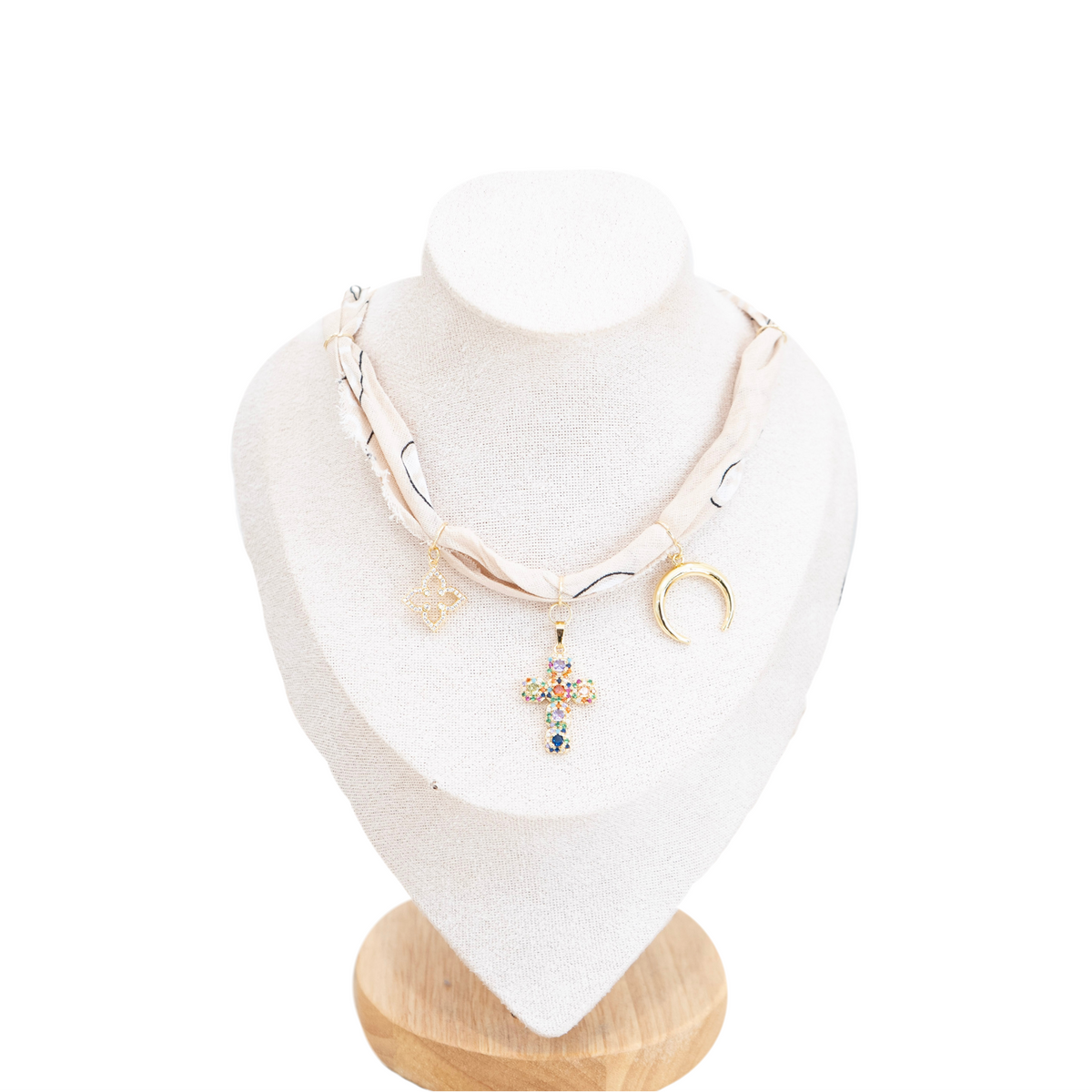 A tan bandana choker-style necklace displayed on a cream fabric jewelry bust. The necklace features a cream-colored printed fabric base with thin black lines, a gold crescent moon charm, a small gold heart charm, and a central gold cross pendant embellished with multicolored rhinestones.