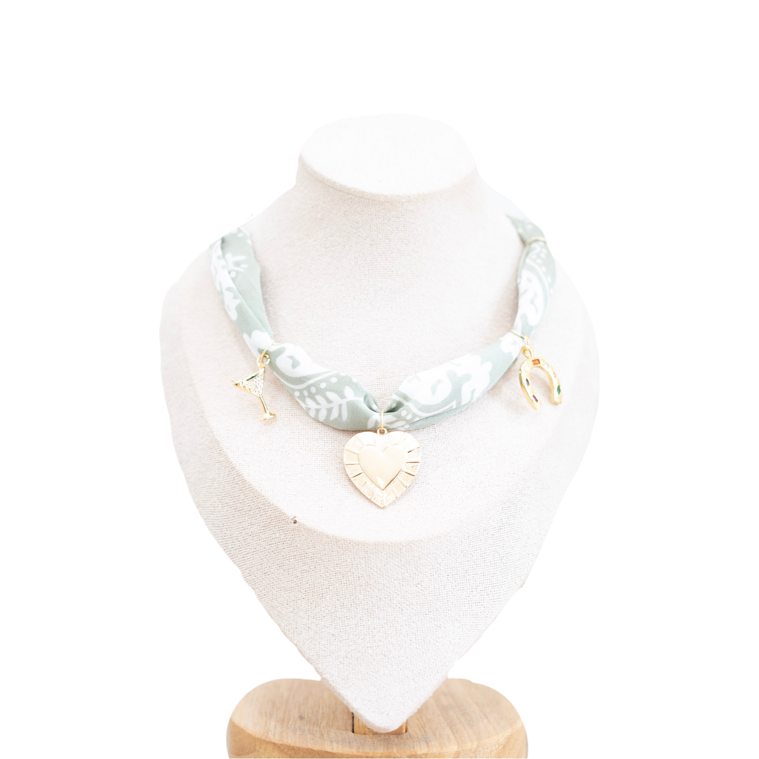 A necklace displayed on a cream jewelry bust against a white background. The necklace features a sage green and white patterned fabric ribbon with gold-toned hardware, including a heart-shaped charm and a small dangling charm.