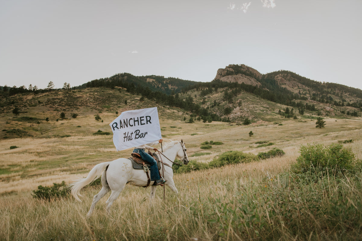 A person riding a white horse across an open grassy landscape with hills and rocky mountains in the background, holding a white sign that reads 'RANCHER Hat Bar'.