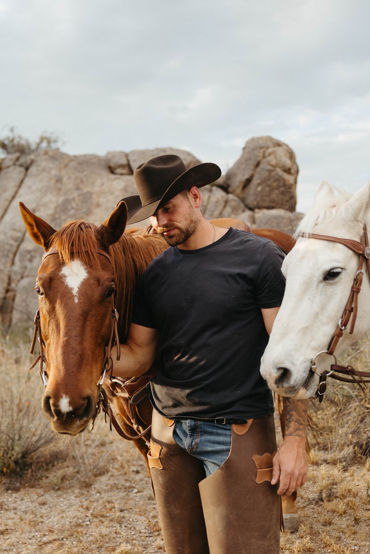 Man in cowboy hat black shirt and chaps with two horses in the desert
