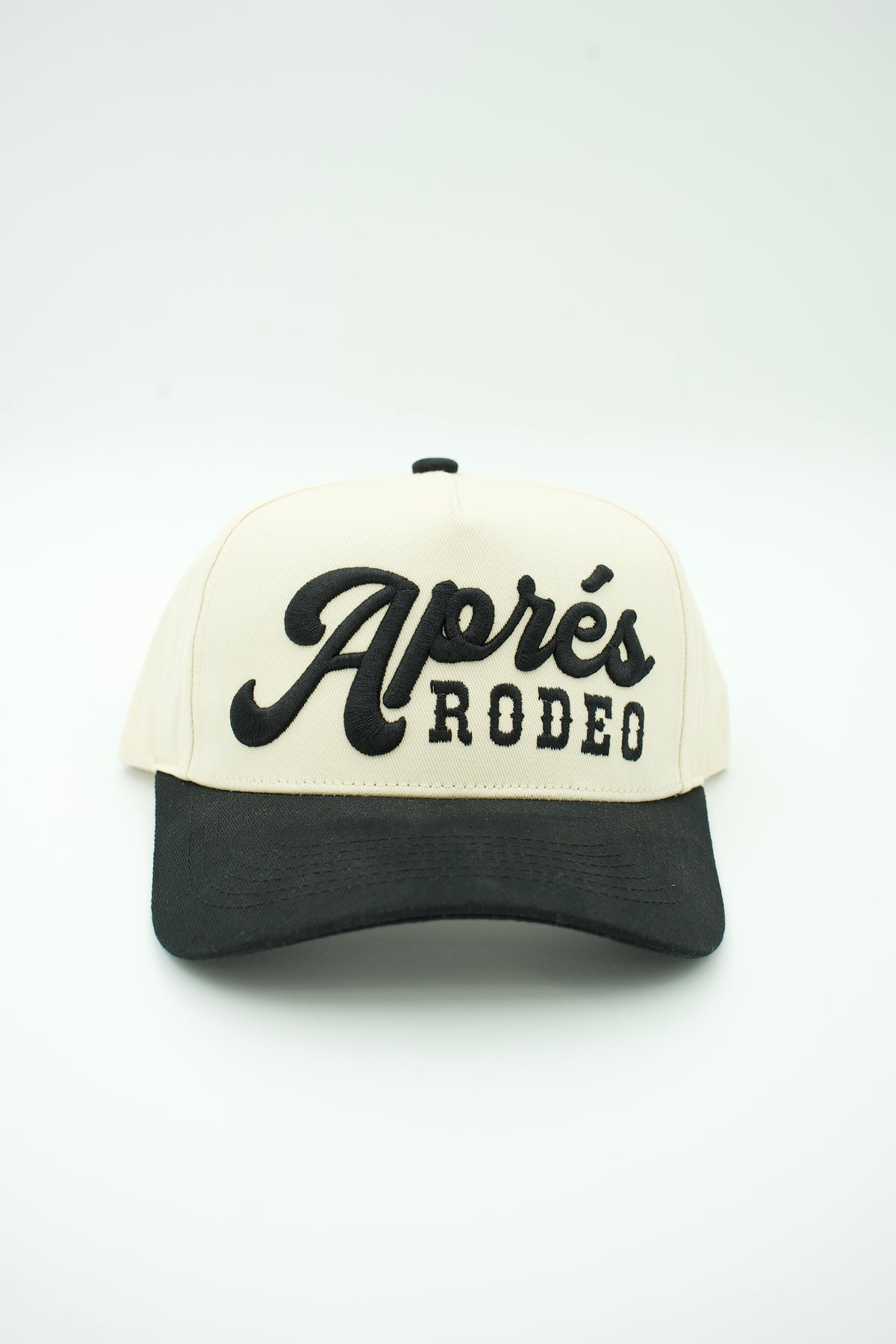 A two-tone baseball cap with an off-white front panel and brim, and a black brim. The front panel features the embroidered text 'Aprés RODEO' in black, with 'RODEO' in block letters and 'Aprés' in a cursive script.