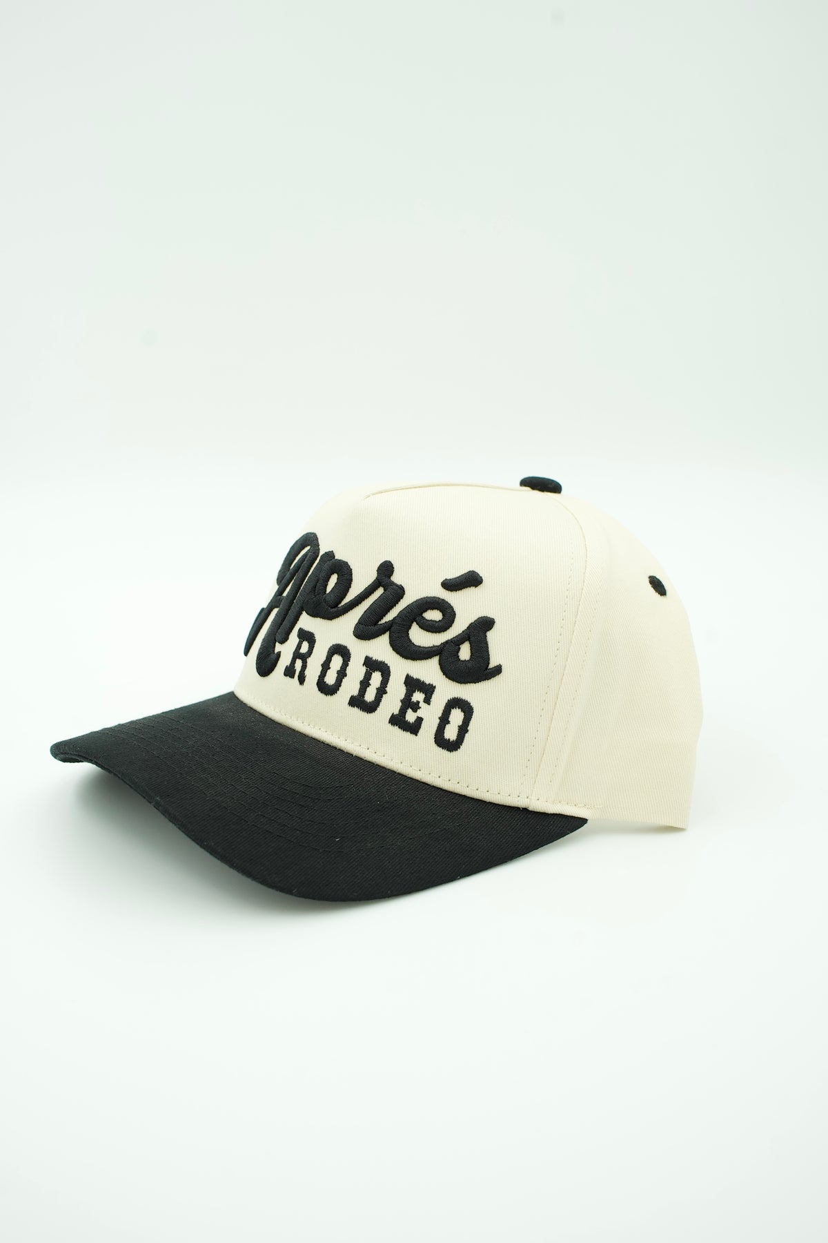 A two-tone baseball cap with an off-white crown and a black brim, featuring black embroidered text that reads 'Aprés RODEO' on the front.
