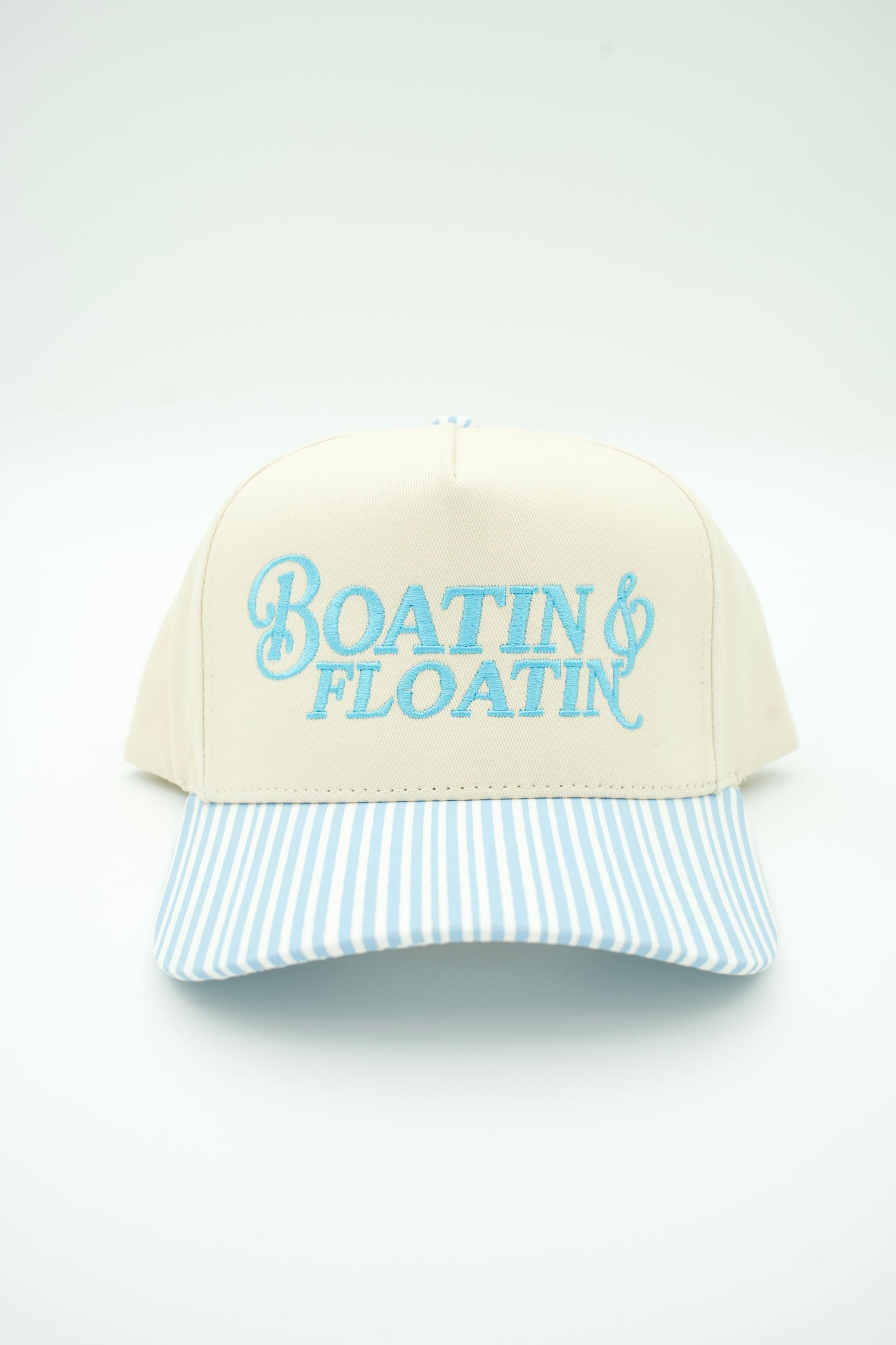 A cream-colored trucker-style cap with light blue embroidered text reading 'Boatin' & Floatin'' on the front, featuring a light blue and white striped brim, set against a plain white background.