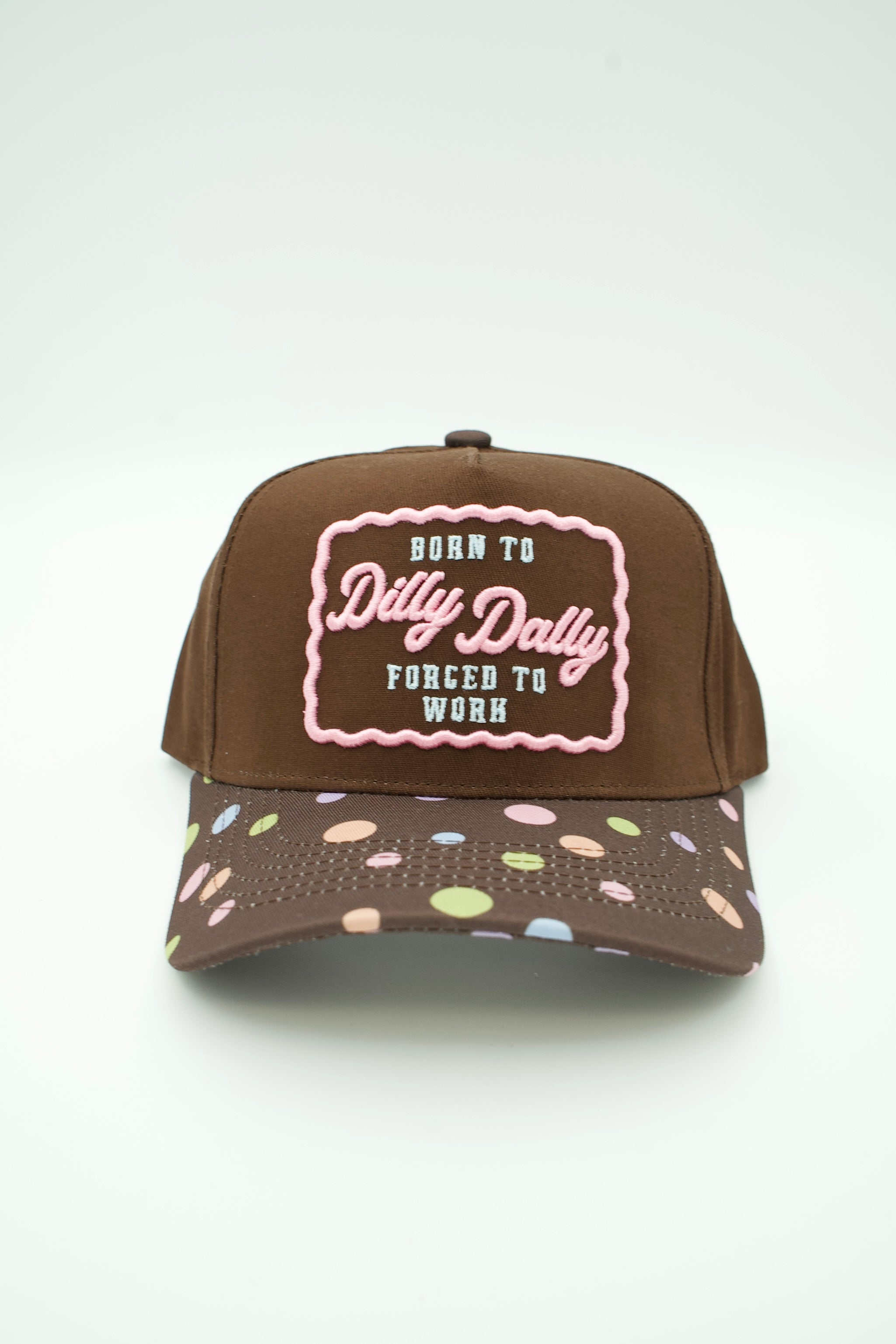 A brown trucker-style cap with a front patch reading 'BORN TO Dilly Dally FORCED TO WORK' in pink and light blue, plus a brim covered in pastel multicolor polka dots.
