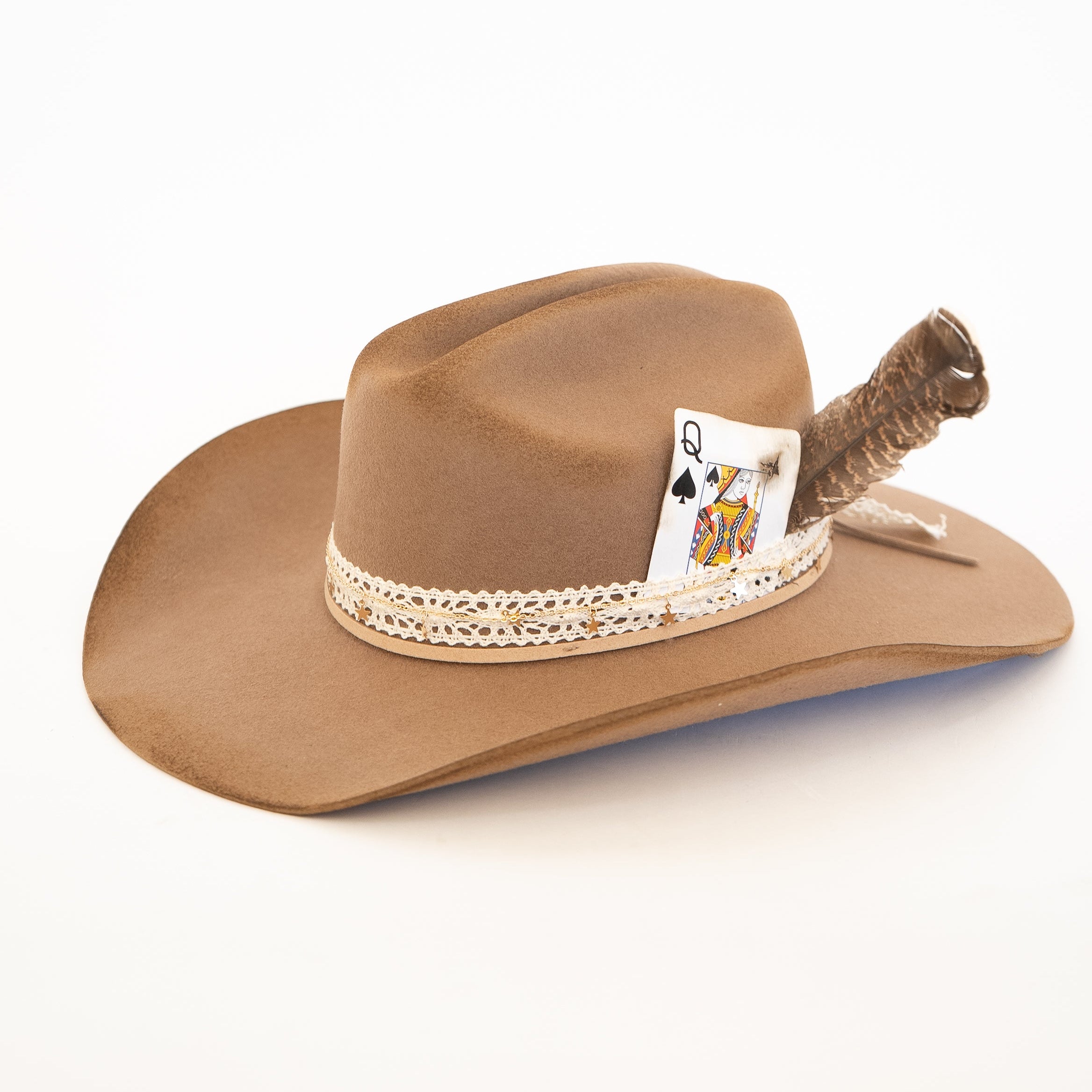 A brown felt cowboy hat with a decorative band featuring a lace trim and a playing card accent, topped with a feather.