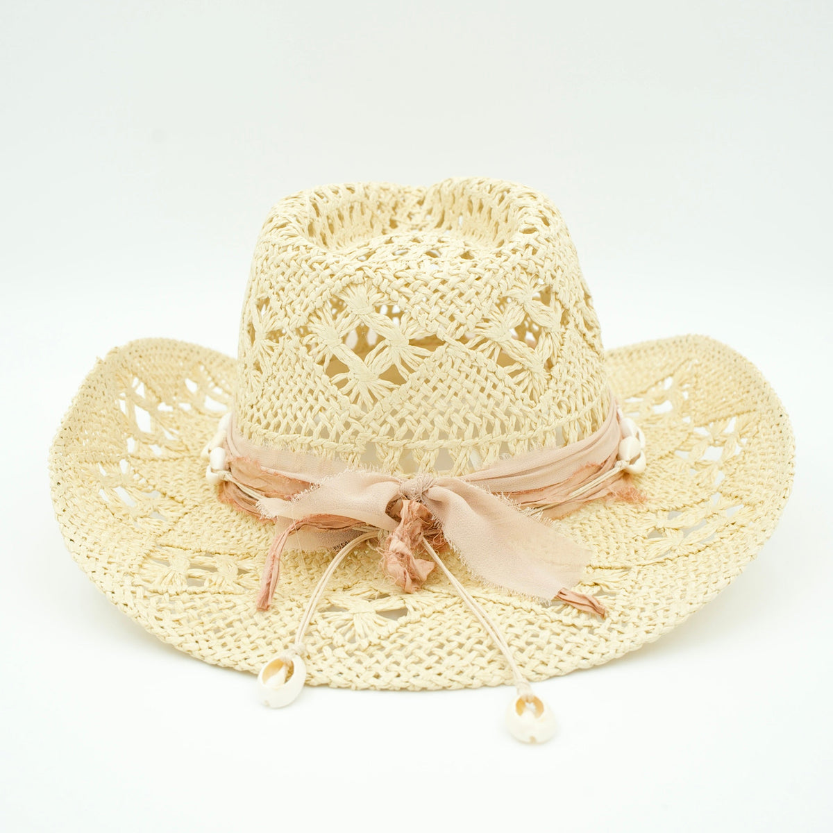 A light beige crocheted cowboy hat with a wide brim, featuring a pink ribbon band tied in a bow and small white shell-like accents hanging from the front.