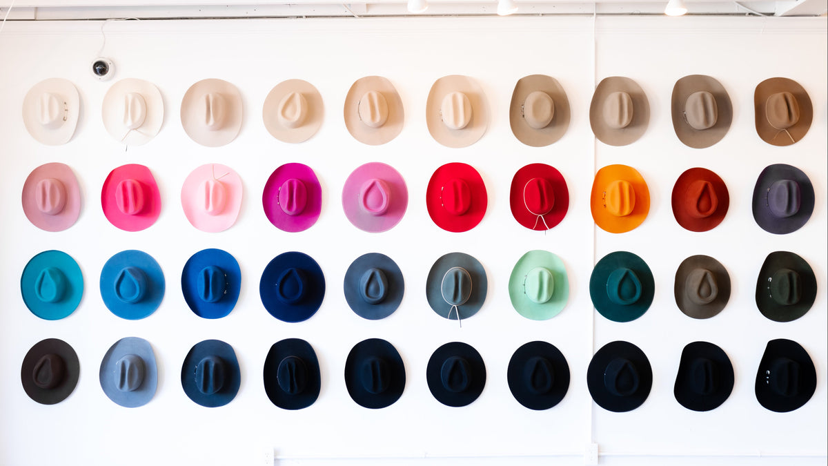 A grid of many felt hats displayed on a white wall, showing a wide range of colors including beige, brown, pink, red, orange, blue, green, and black.