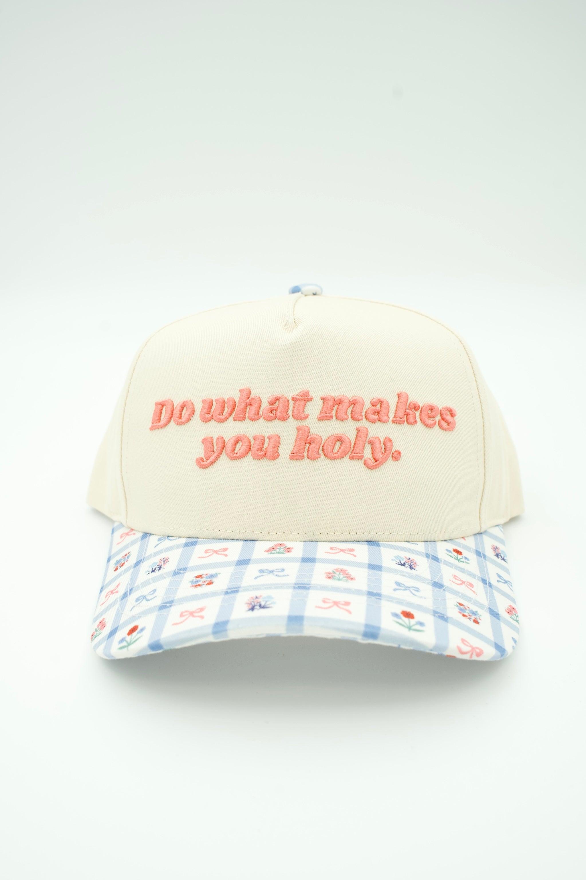 An off-white baseball cap with orange embroidered text reading 'do what makes you holy.' on the front, featuring a light blue and white gingham brim with small embroidered floral motifs.