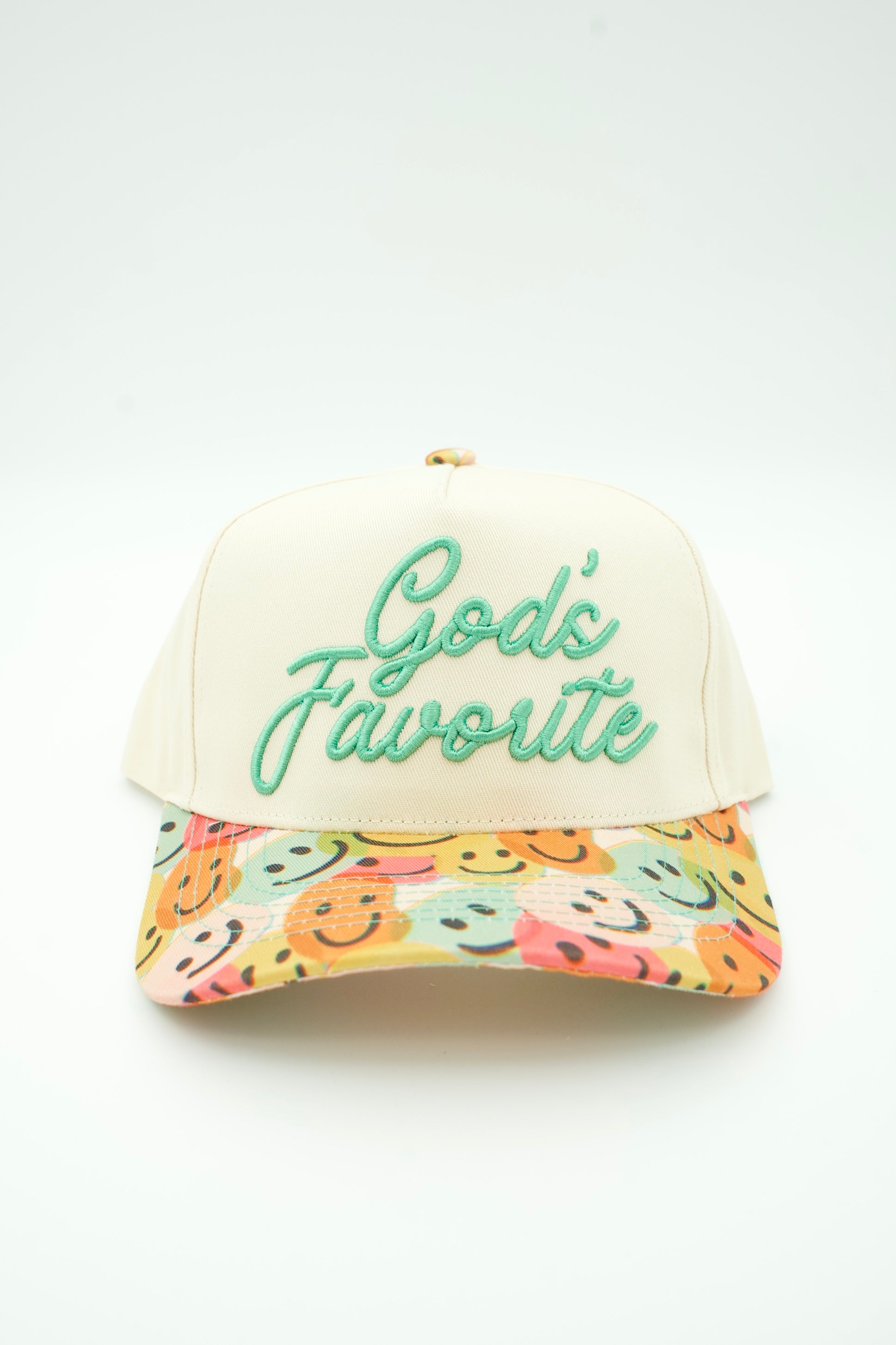A baseball cap with an off-white crown and a colorful patterned brim featuring smiley faces. The front has green embroidered text reading 'God's Favorite'.