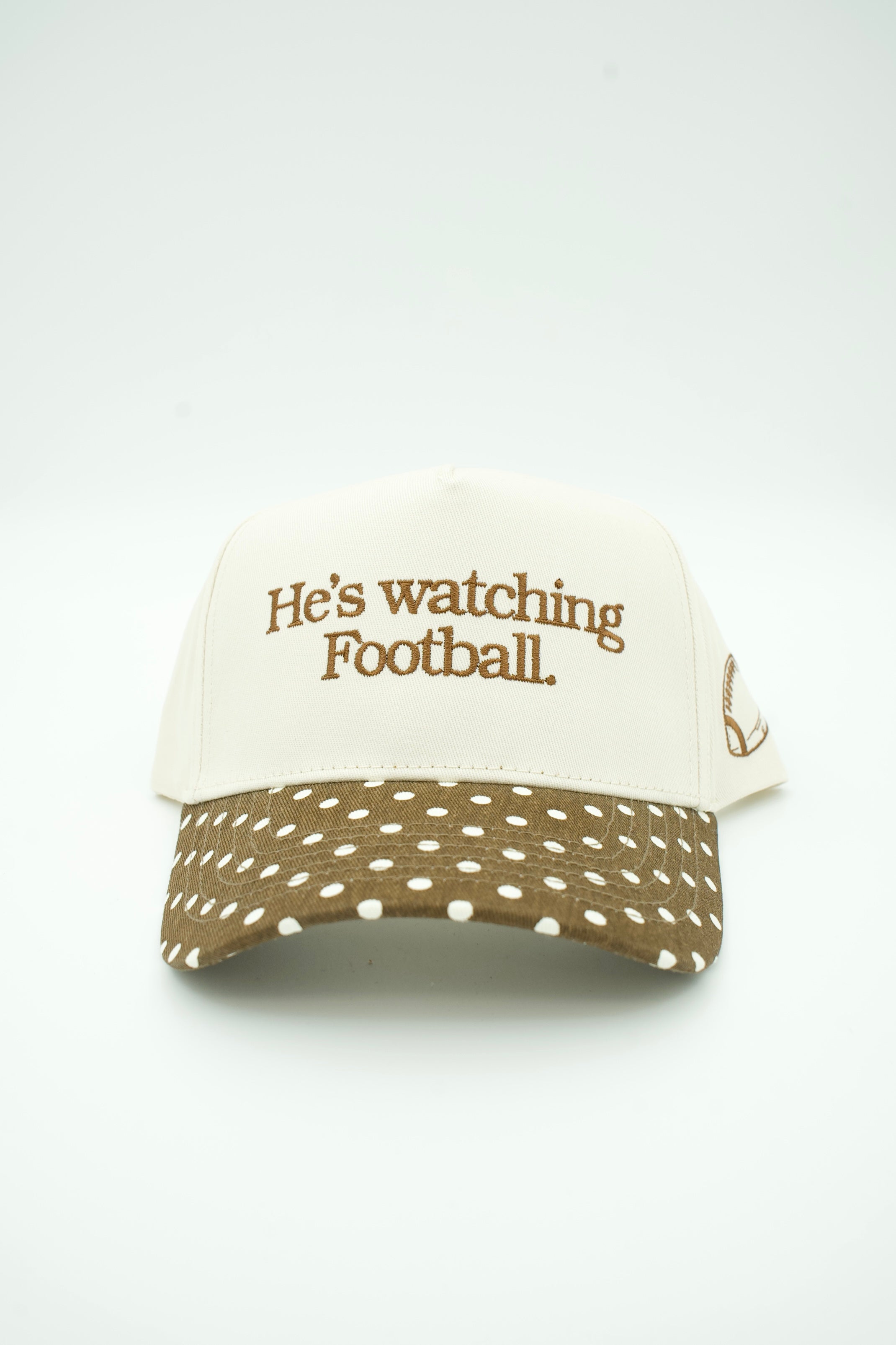 A baseball cap with an off-white crown and a brown visor covered in white polka dots. The front of the cap features the embroidered brown text 'He's watching Football.
