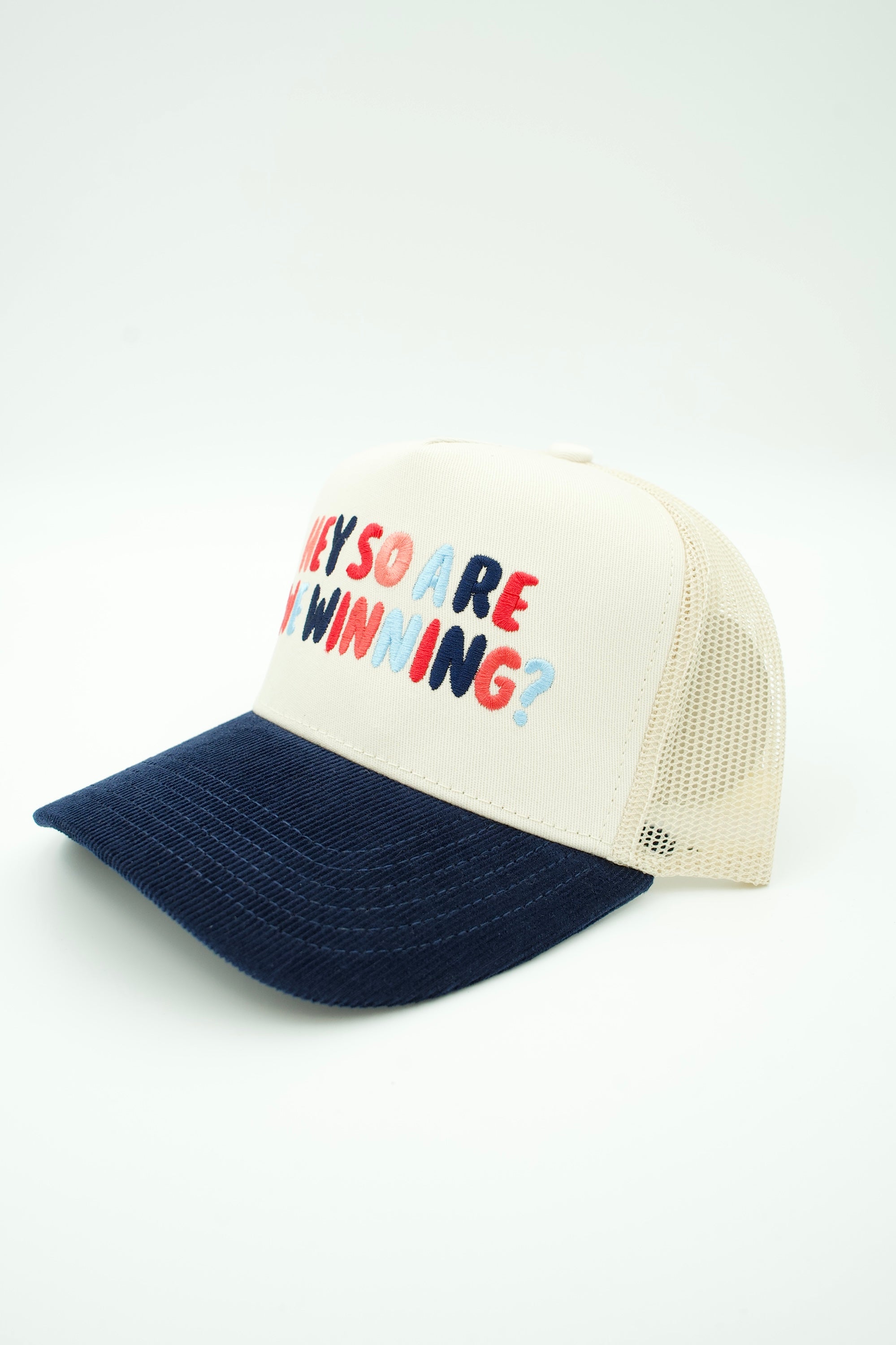 A cream and navy blue trucker hat with the text 'THEY SO ARE WE WINNING?' embroidered on the front in red, navy, and light blue.