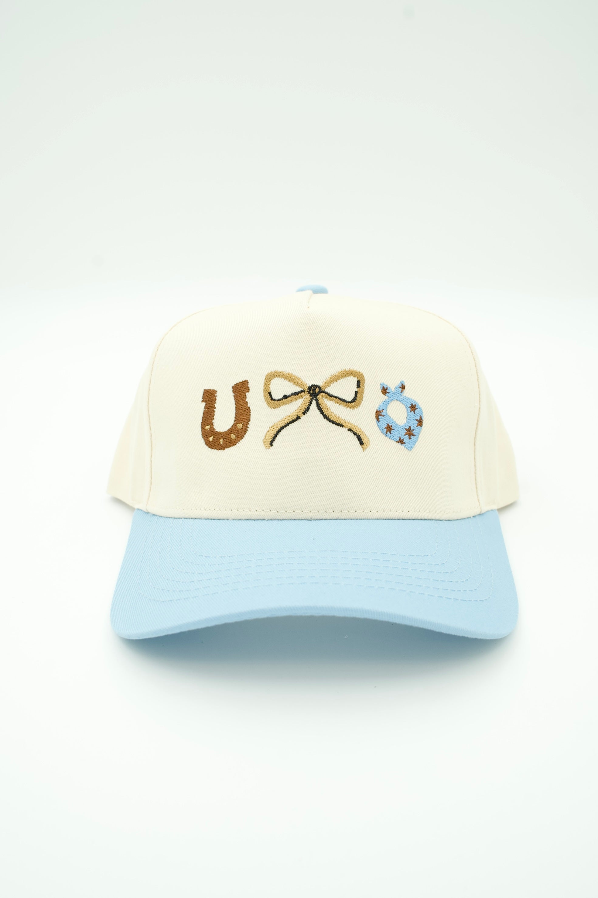 A two-tone baseball cap with an off-white crown and a light blue brim, featuring embroidered horse-themed symbols on the front: a brown horseshoe, a tan bow, and a blue patterned bandana.