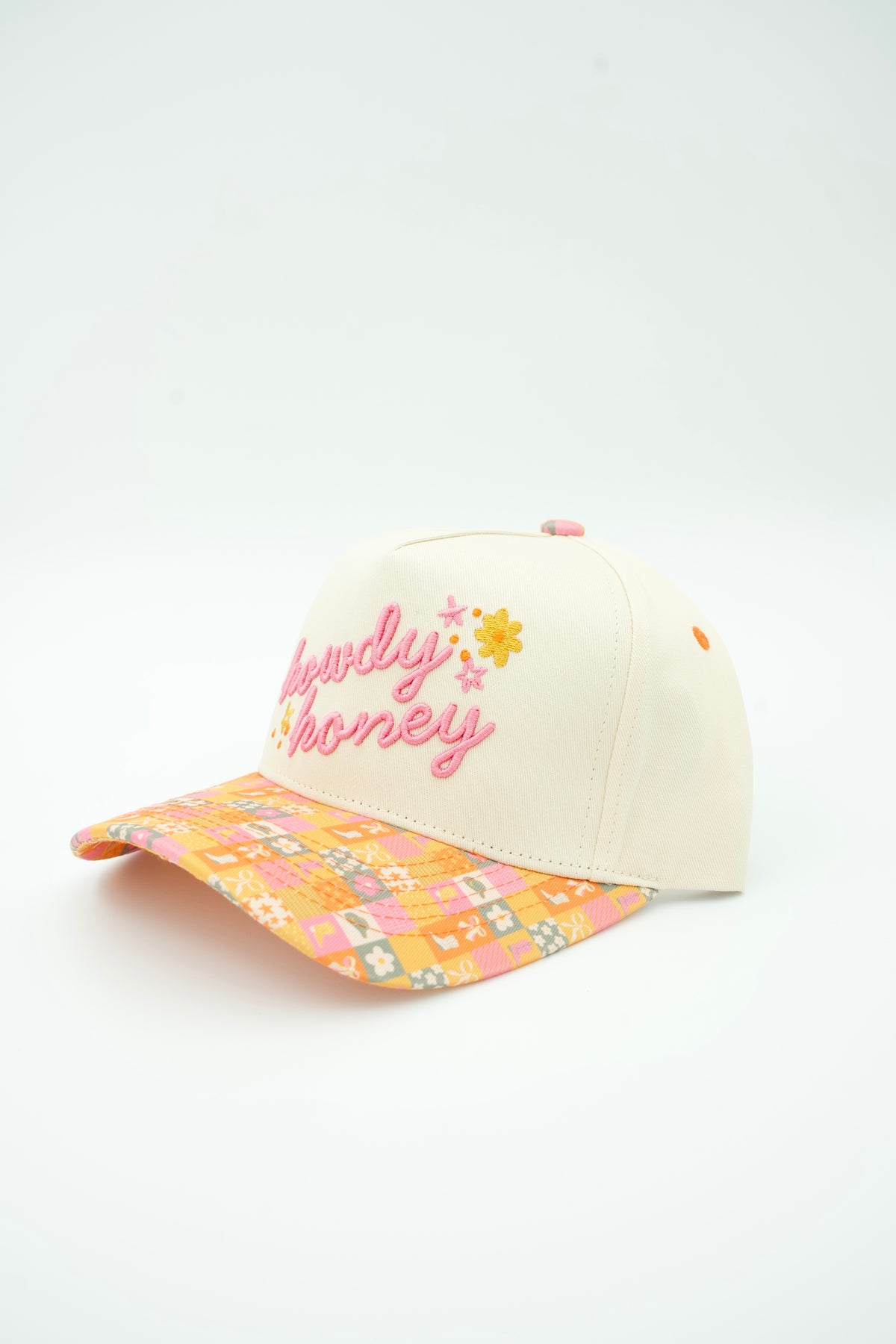 A cream-colored baseball cap with pink embroidered text reading 'honey' and small floral details on the front, featuring a vibrant orange, pink, and yellow patterned brim.