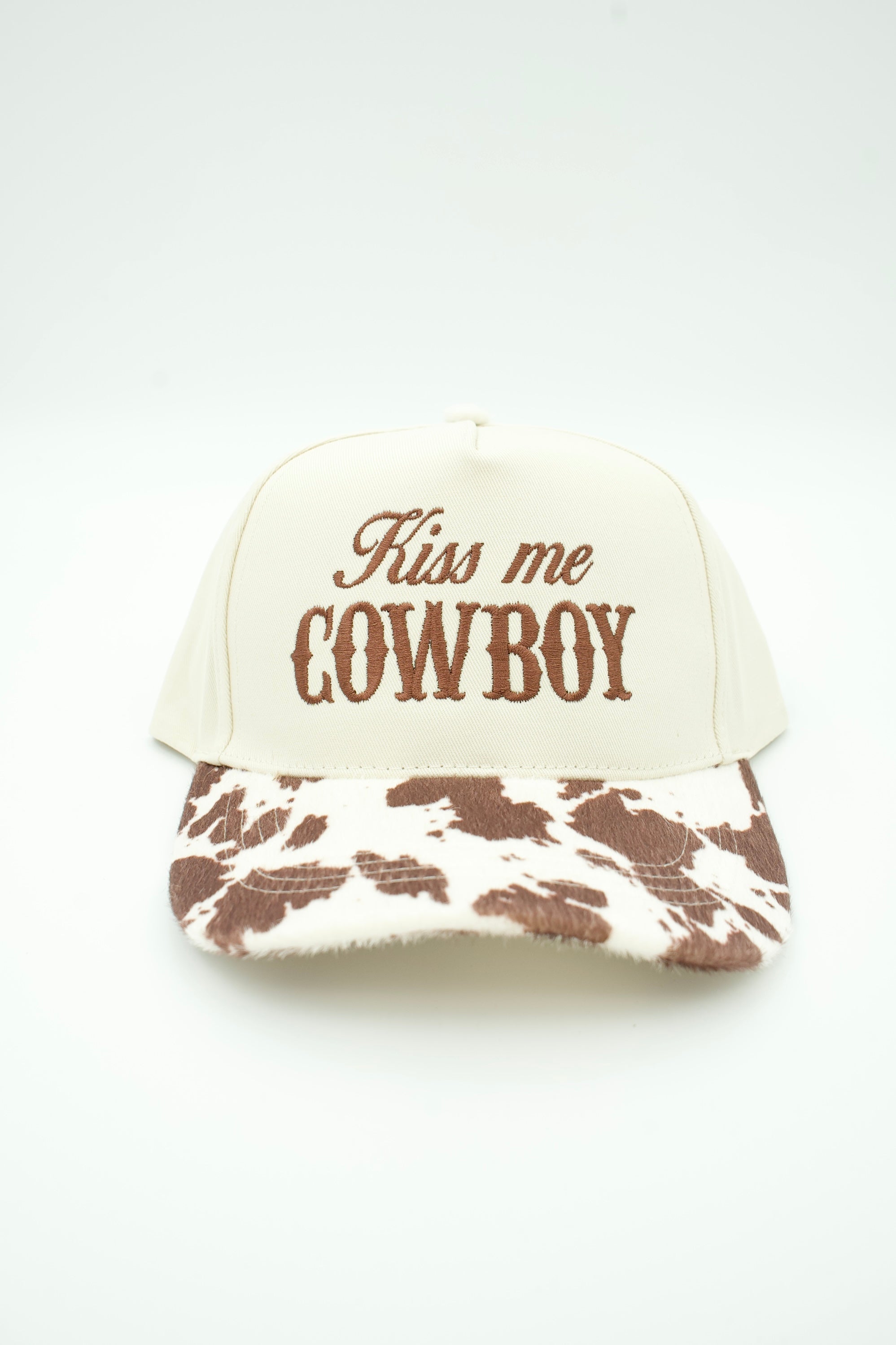 An off-white trucker-style cap with brown embroidered text reading 'Kiss me COWBOY' on the front and a brown-and-white cow-print pattern on the brim, shown against a plain white background.