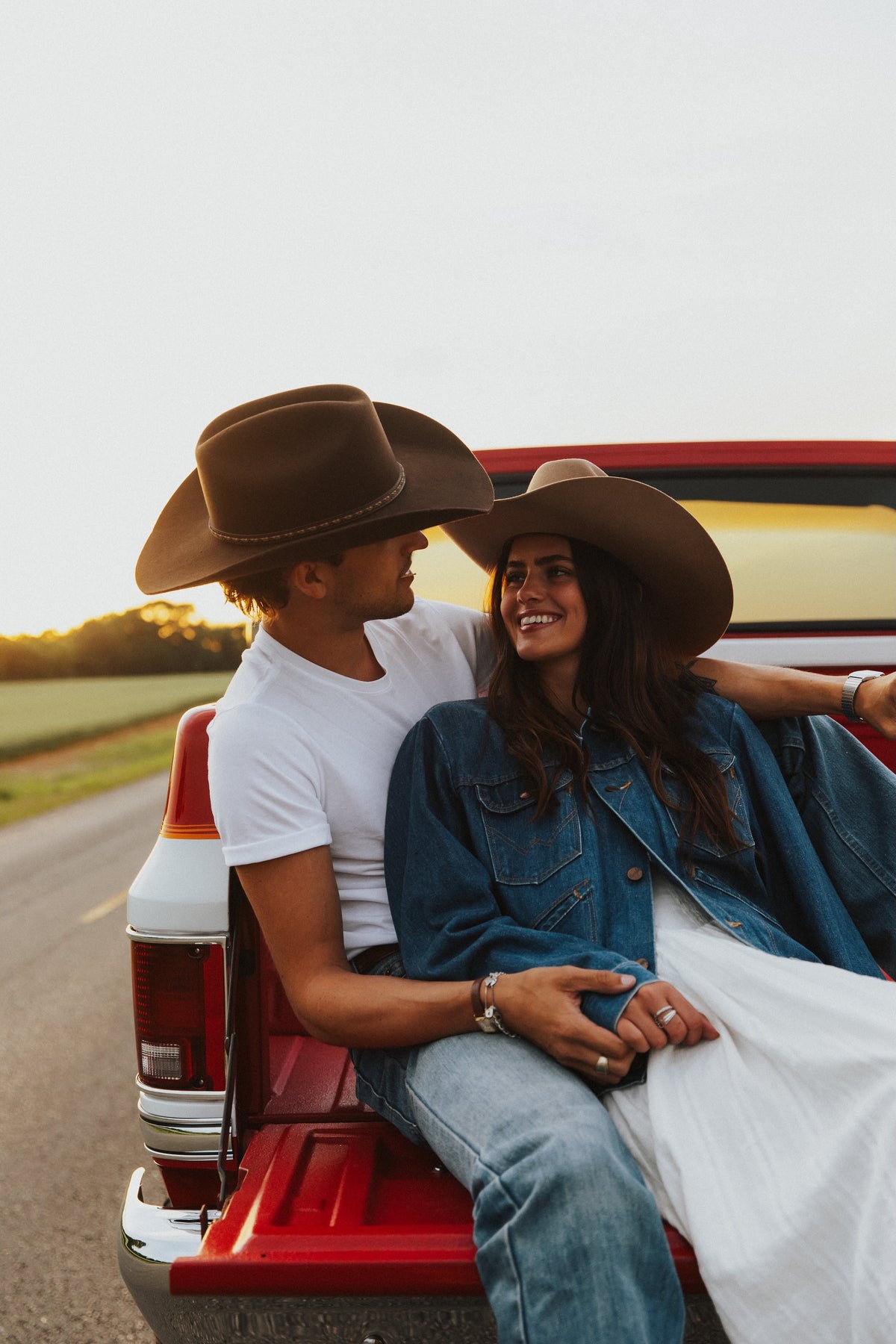 A couple wearing cowboy hats sits in the bed of a red pickup truck on a country road at sunset. The man wears a white t-shirt and jeans, and the woman wears a denim jacket over a white dress.