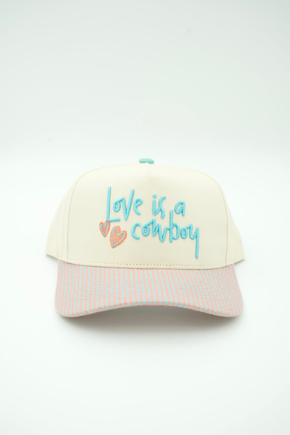 A cream-colored baseball cap with a pastel pink and white striped brim. The front panel features the embroidered text 'love is a cowboy' in light blue, accompanied by two small pink hearts.