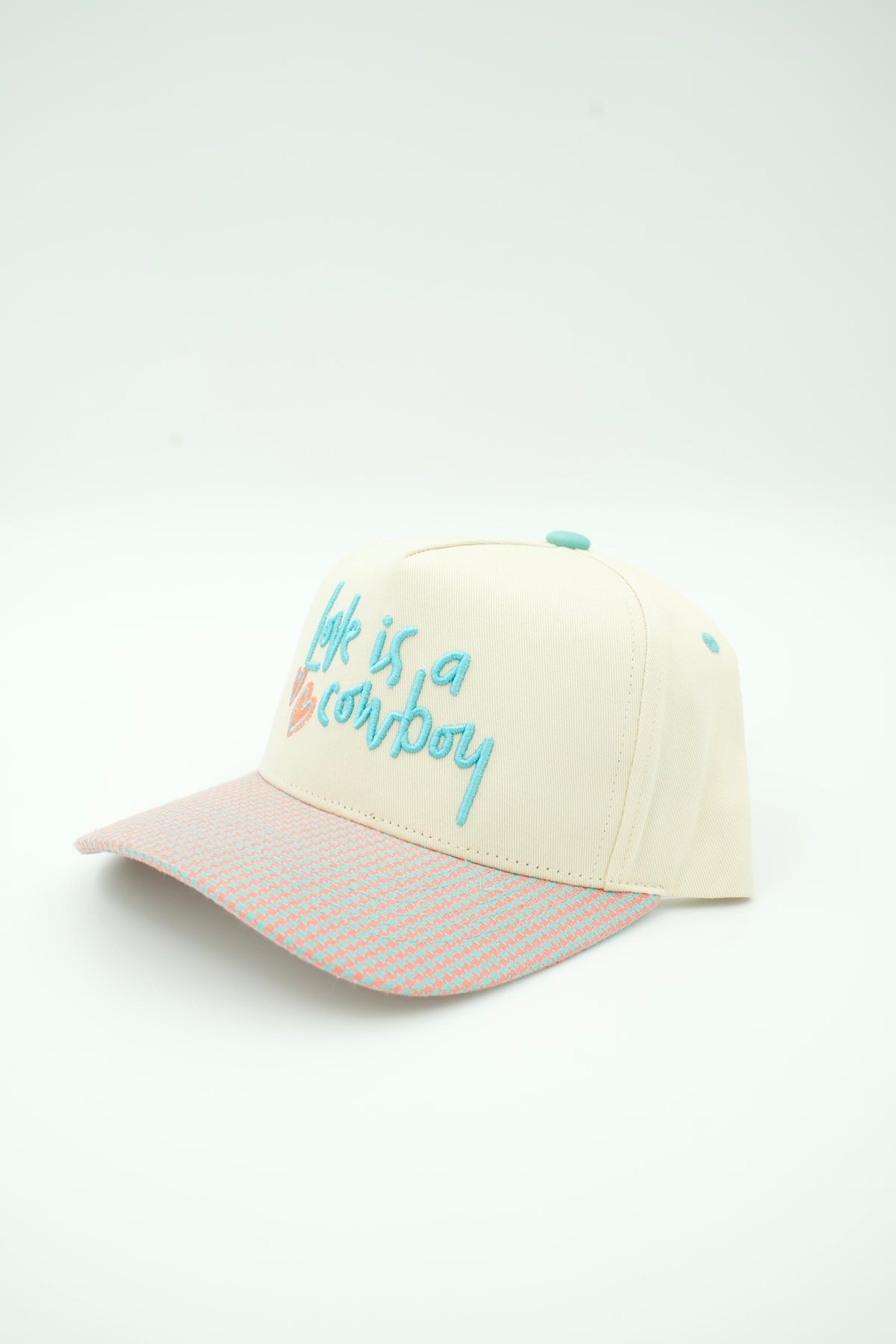 An off-white baseball cap with a patterned light pink brim, featuring embroidered text on the front that reads 'life is a cowboy' in light blue and orange, with a small turquoise button on top.
