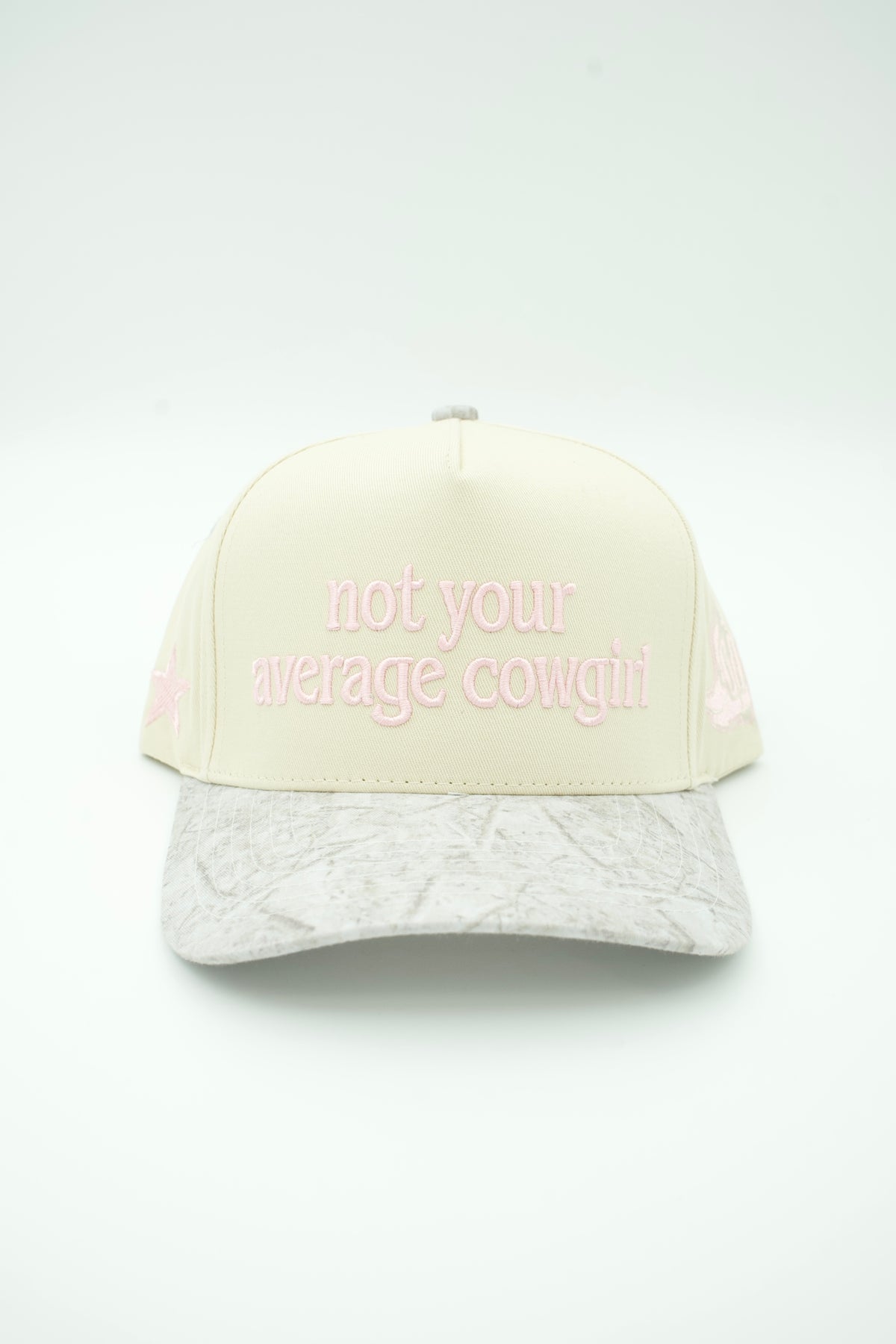A cream-colored trucker-style cap with pink embroidered text reading 'not your average cowgirl' on the front, paired with a light gray crinkled brim and pink side details, set against a plain white background.