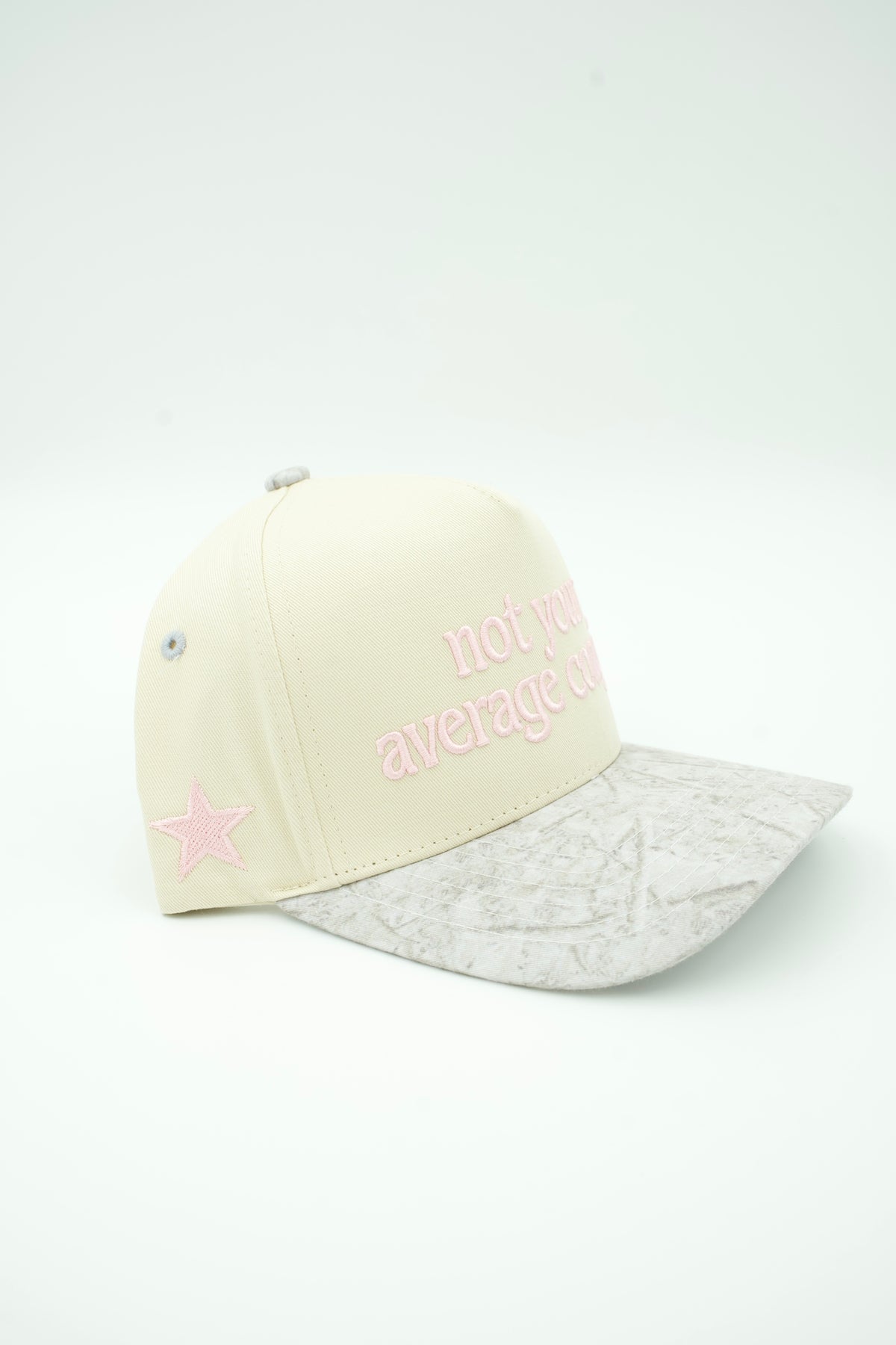 A cream-colored baseball cap with a light gray patterned visor, featuring pink embroidered text that reads 'not an average girl' and a pink star on the side.