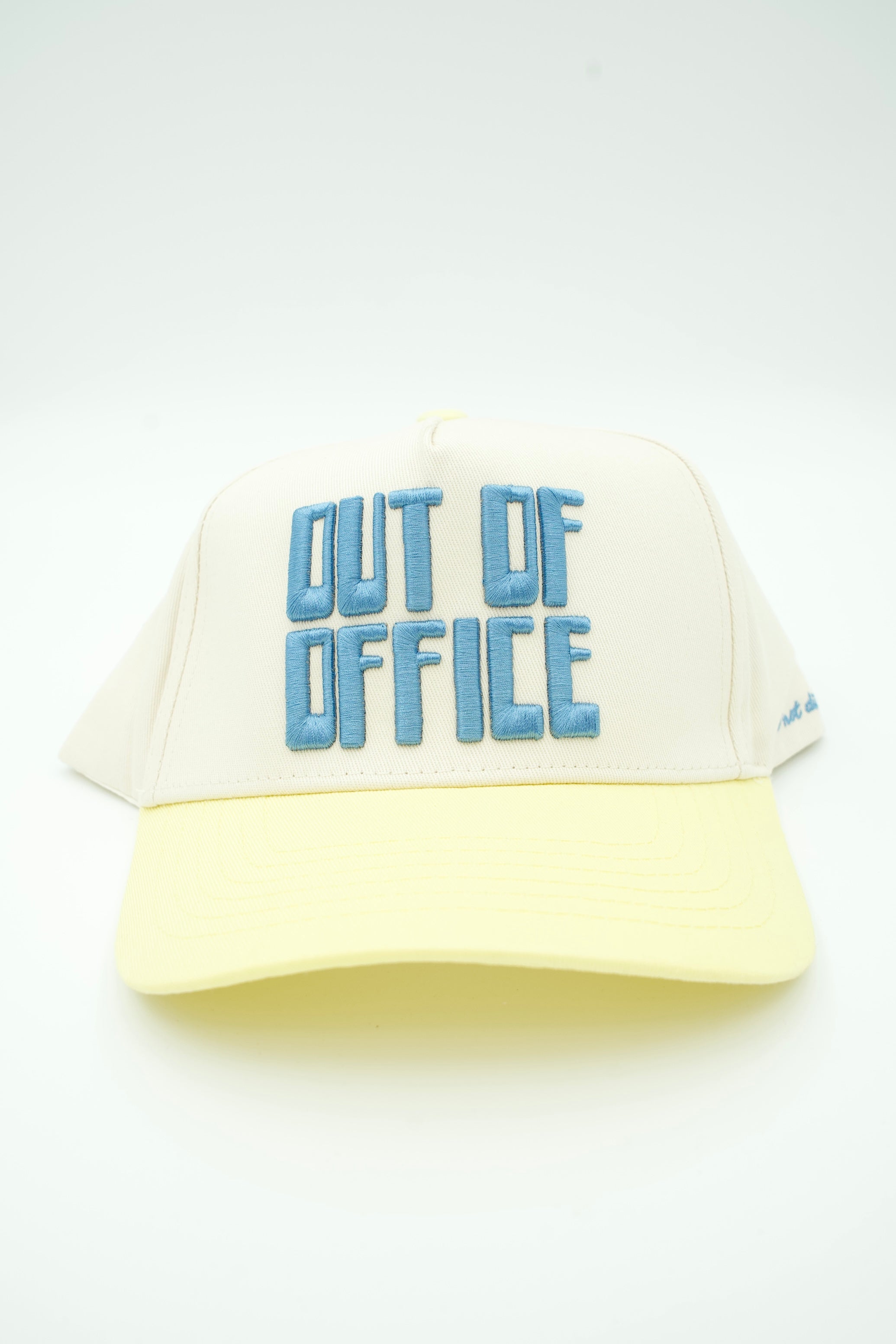 A cream-colored trucker hat with a pale yellow visor and light blue embroidered text reading 'OUT OF OFFICE' on the front, set against a plain white background.