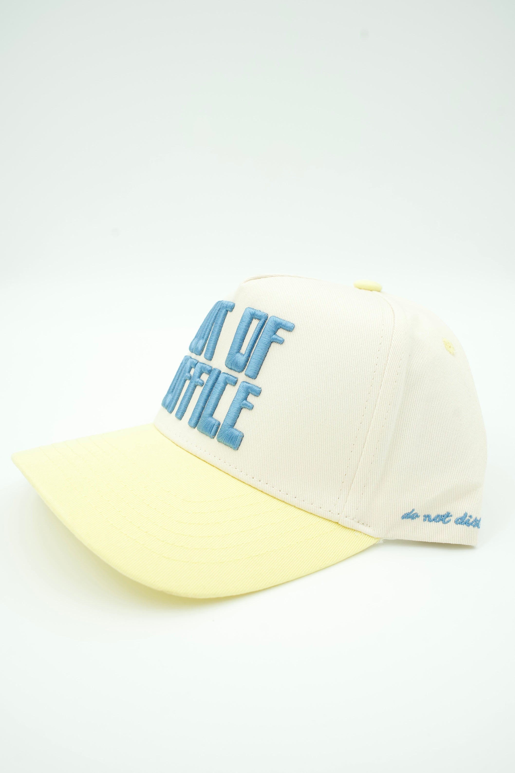 A baseball cap with an off-white crown and a pale yellow brim, featuring light blue embroidered text reading 'OUT OF OFFICE' on the front and 'do not dist' on the side.