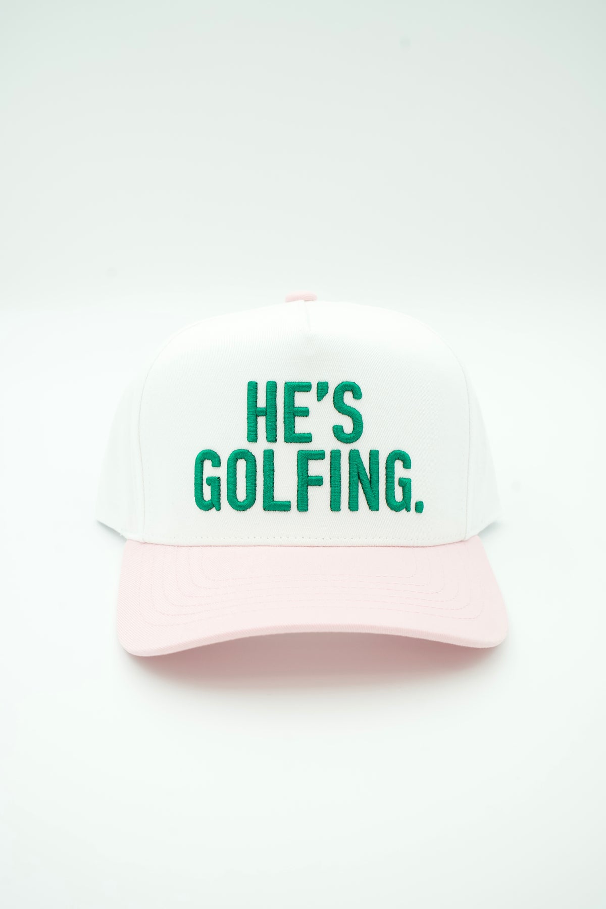 A white baseball cap with a light pink curved brim and green embroidered text reading 'HE'S GOLFING.' on the front, shown against a plain white background.
