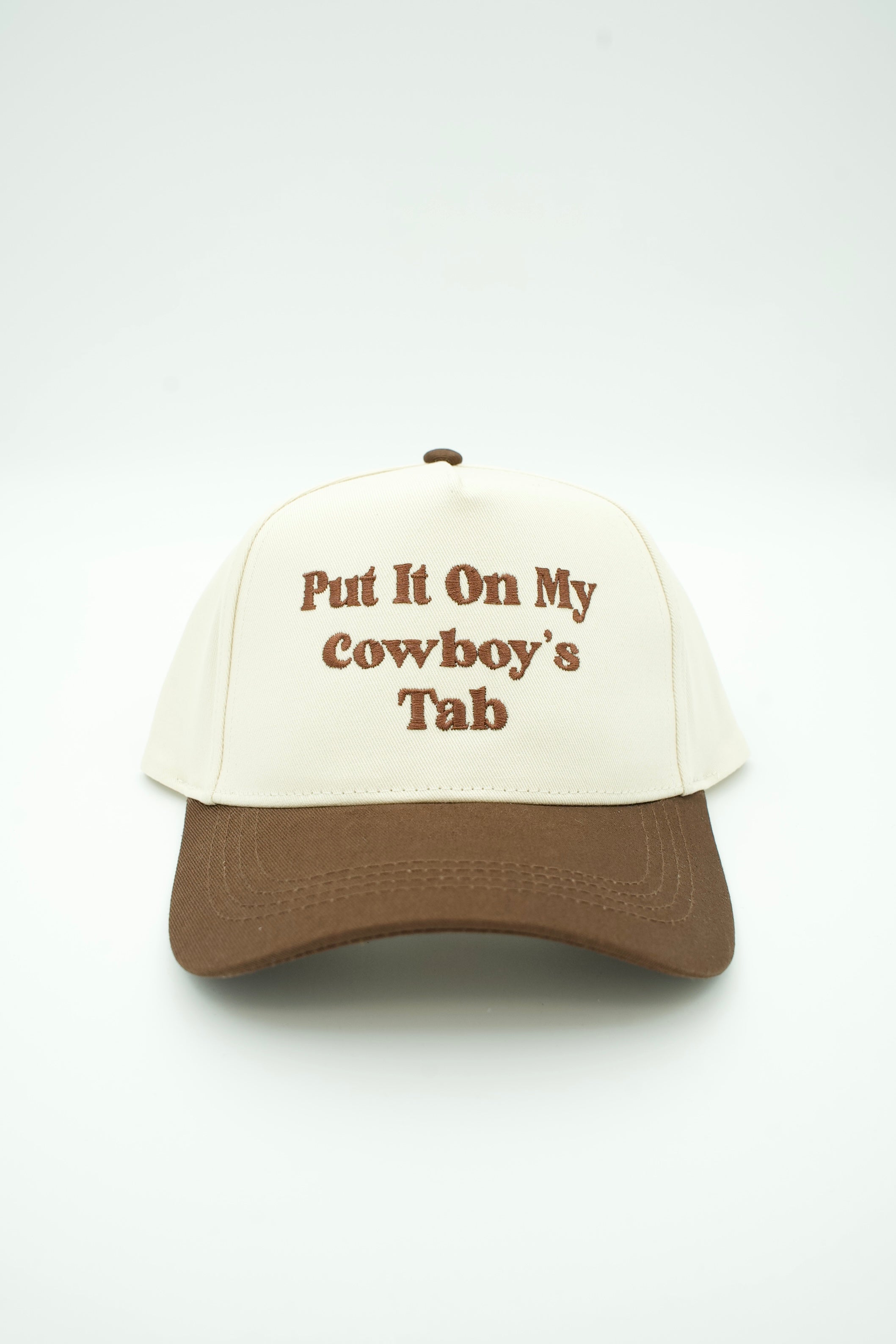 A baseball cap with an off-white crown and a brown visor, featuring the text 'Put It On My Cowboy's Tab' embroidered in brown on the front.