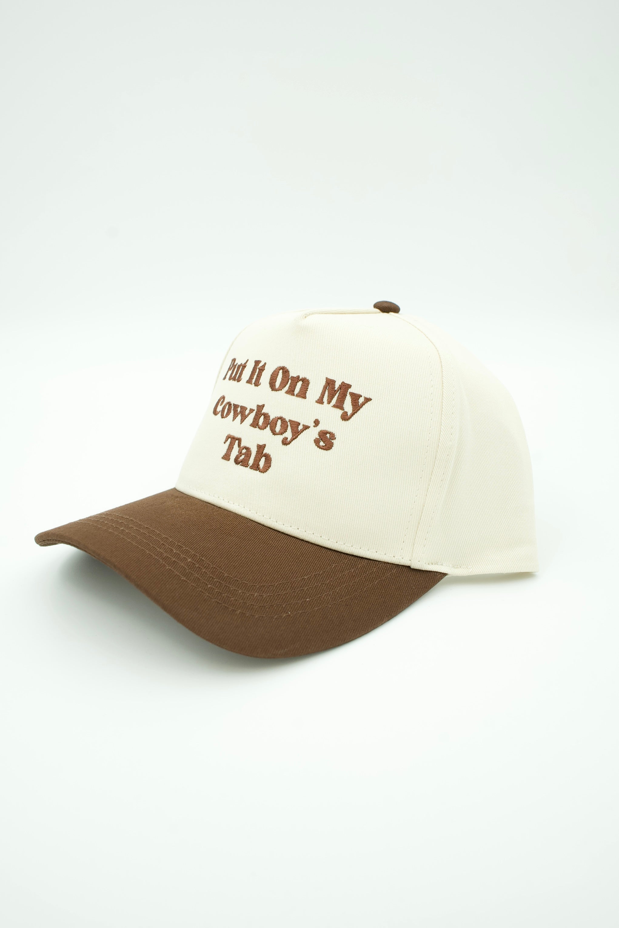 A baseball cap with an off-white crown and a brown visor, featuring brown embroidered text on the front that reads 'Put It On My Cowboy's Tab'.