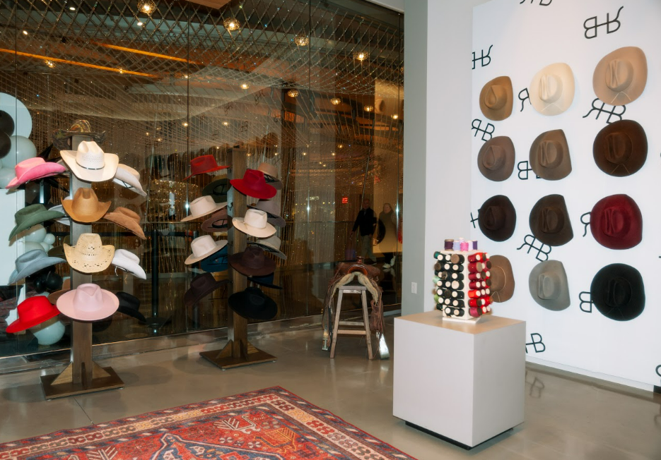 Rancher Hat Bar Las Vegas interior image of a retail display of cowboy and wide-brim hats inside a store. Several hats are shown on wooden display stands near a glass window, while a white wall on the right features multiple circular hat samples in different colors.