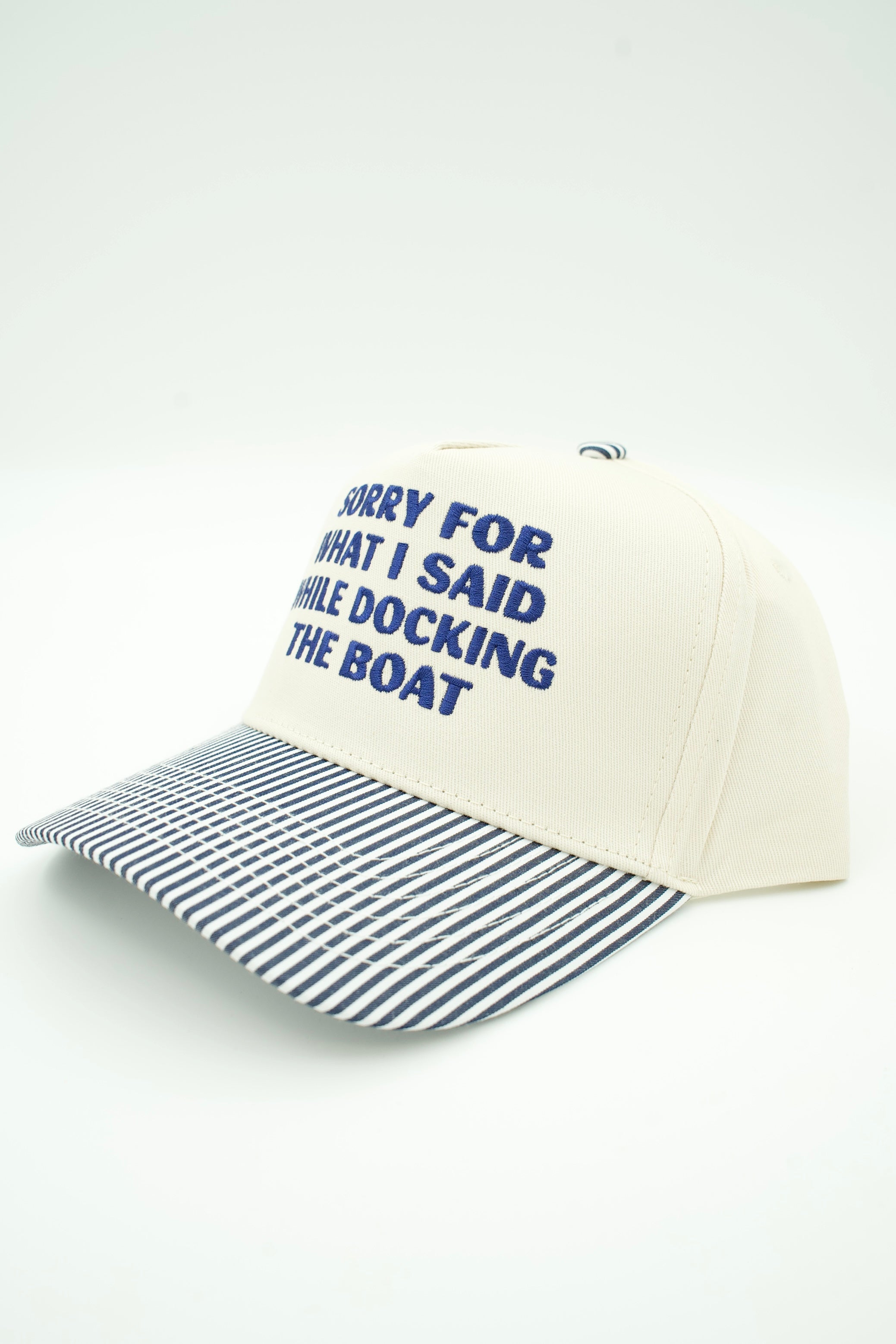 An off-white baseball cap with blue text reading 'SORRY FOR WHAT I SAID WHILE DOCKING THE BOAT' on the front panel and a navy-and-white striped brim, shown on a clean white background.