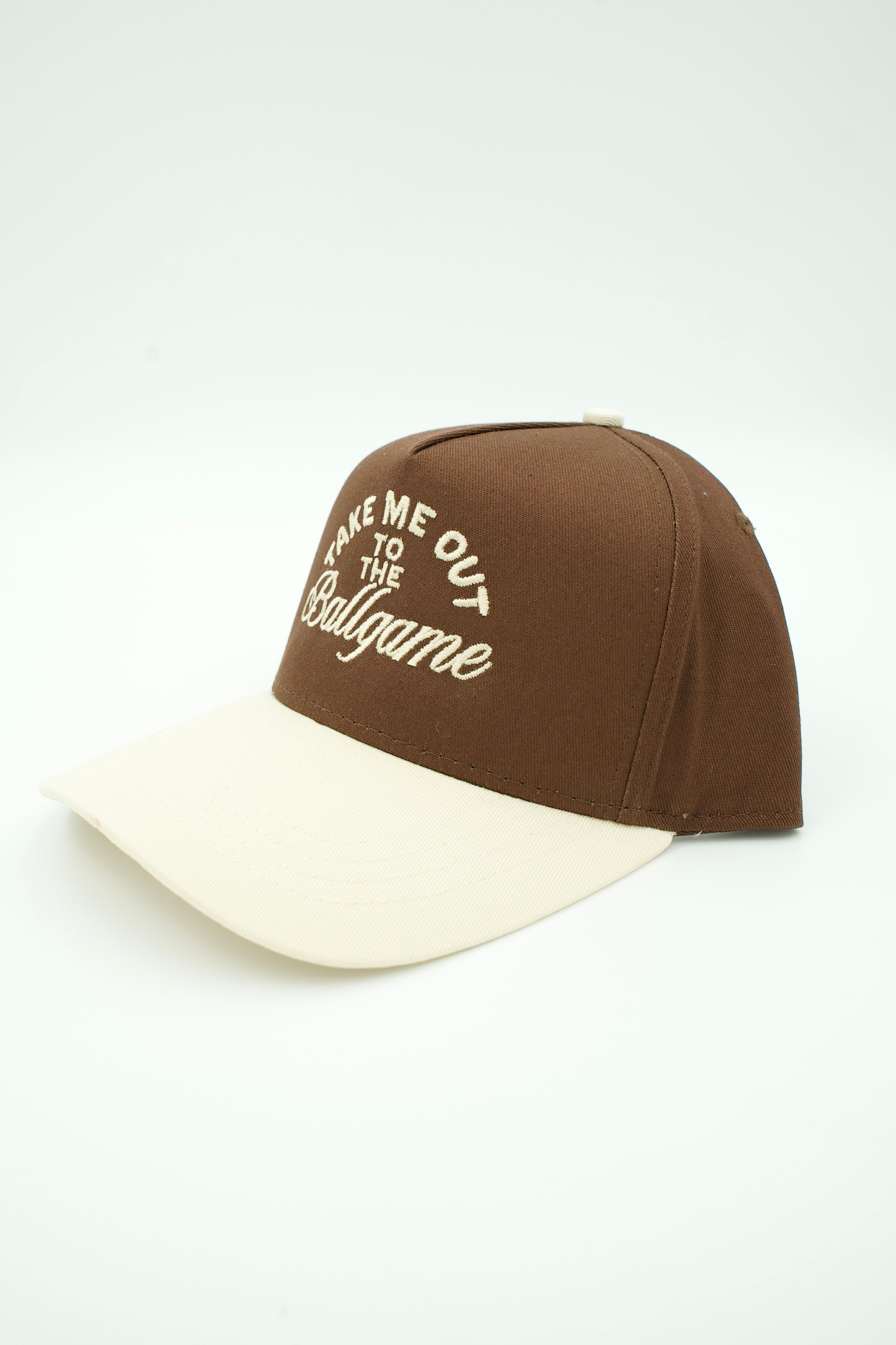 A two-tone baseball cap with a brown crown and a cream-colored visor. The front of the crown features white embroidered text that reads 'TAKE ME OUT TO THE Ballgame'.