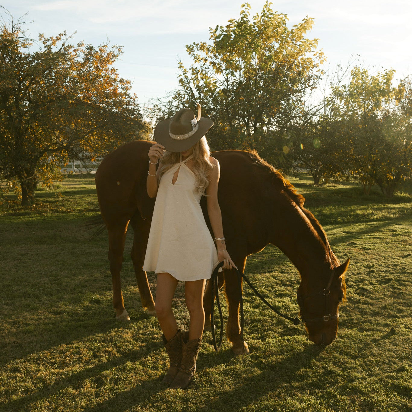 Woman in a white dress standing next to a horse in a field with trees in the background