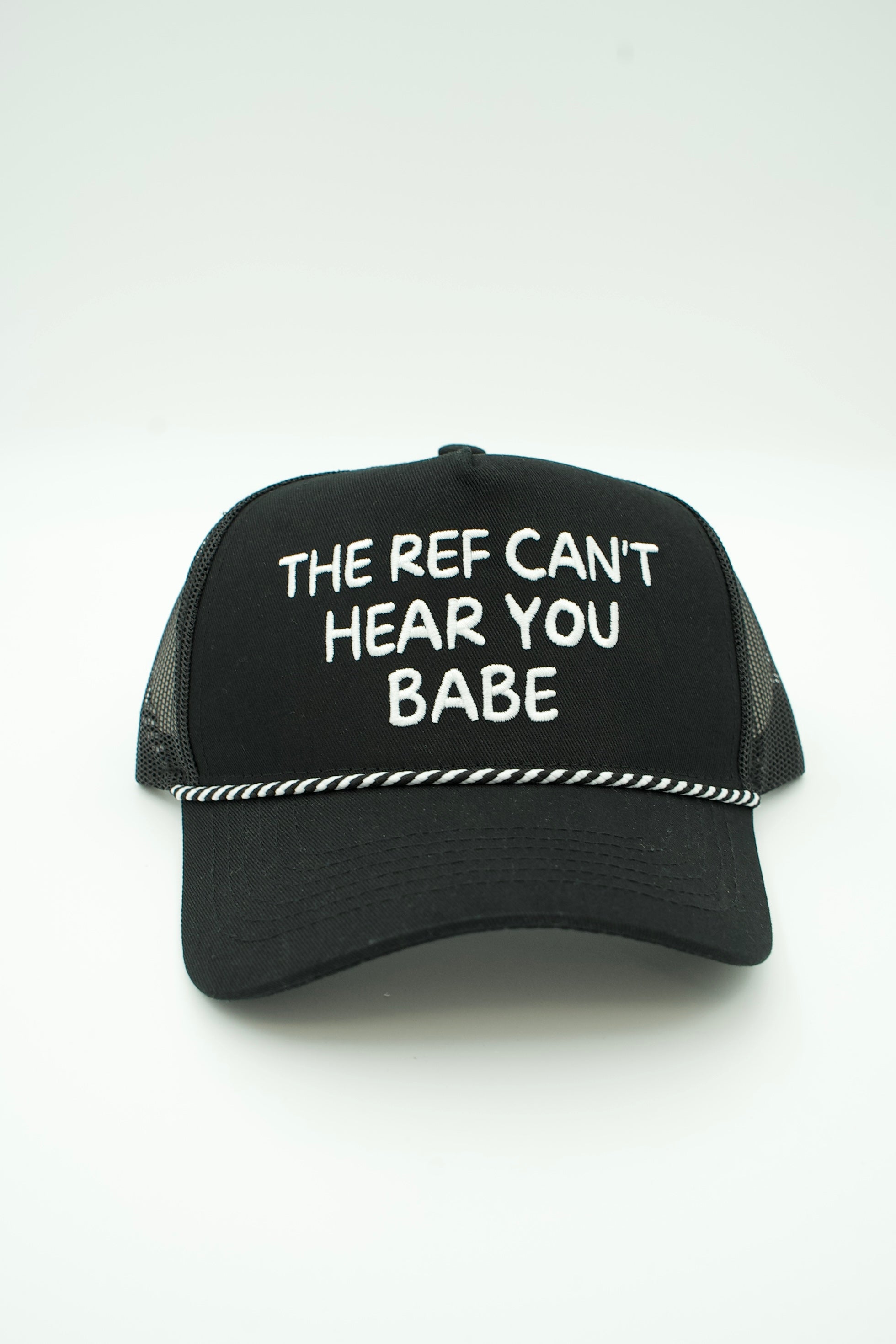 A black trucker-style baseball cap with white embroidered text reading 'THE REF CAN'T HEAR YOU BABE' on the front, shown against a plain white background.