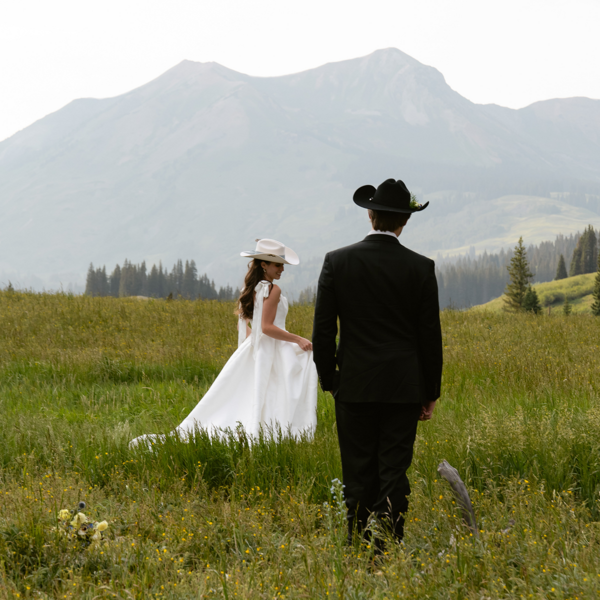 A bride in a white wedding dress and hat and a groom in a black suit and cowboy hat standing in a grassy field with mountains in the background.