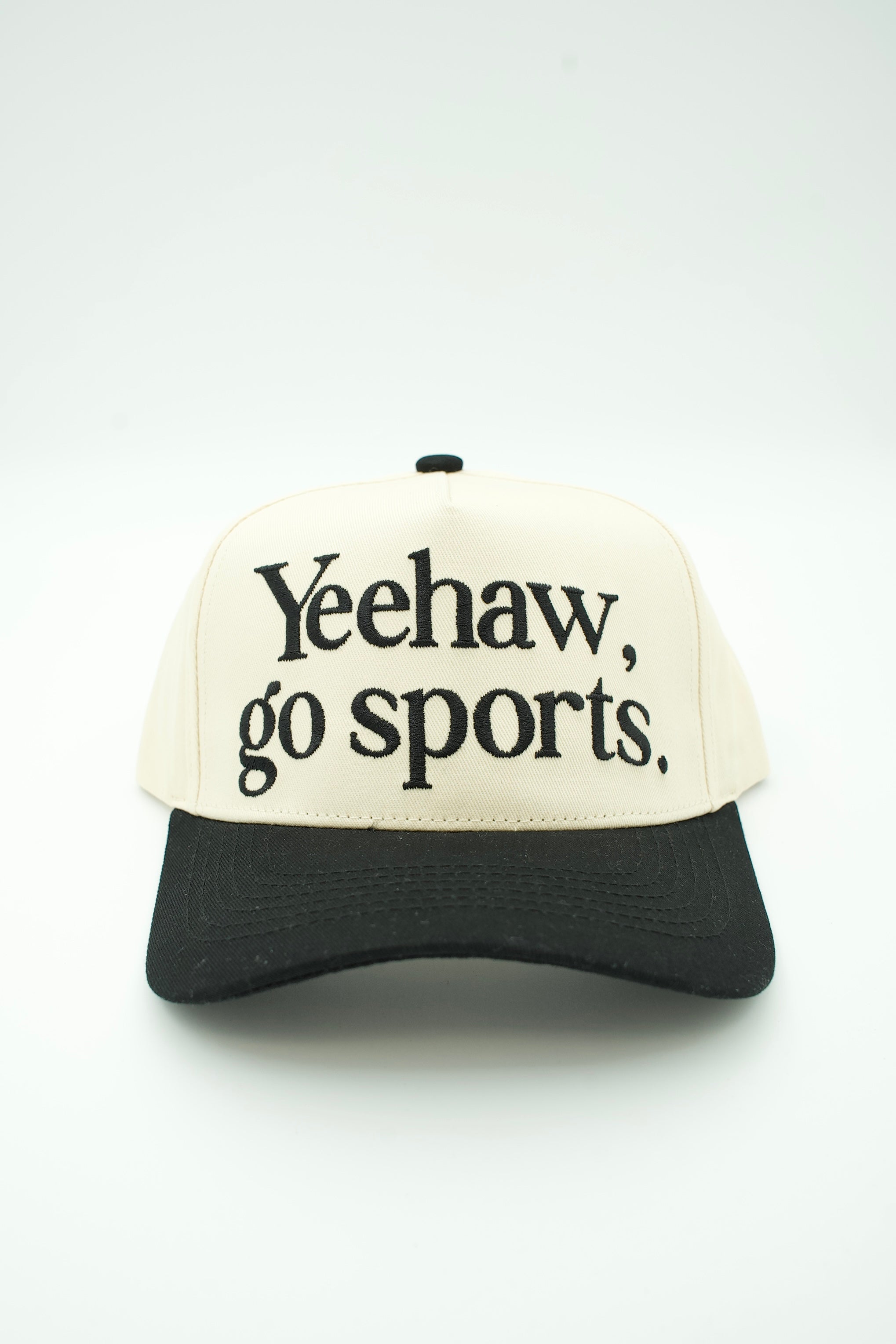 A two-tone baseball cap with an off-white front panel and black brim, featuring the text 'Yeehaw, go sports.' embroidered in black on the front.