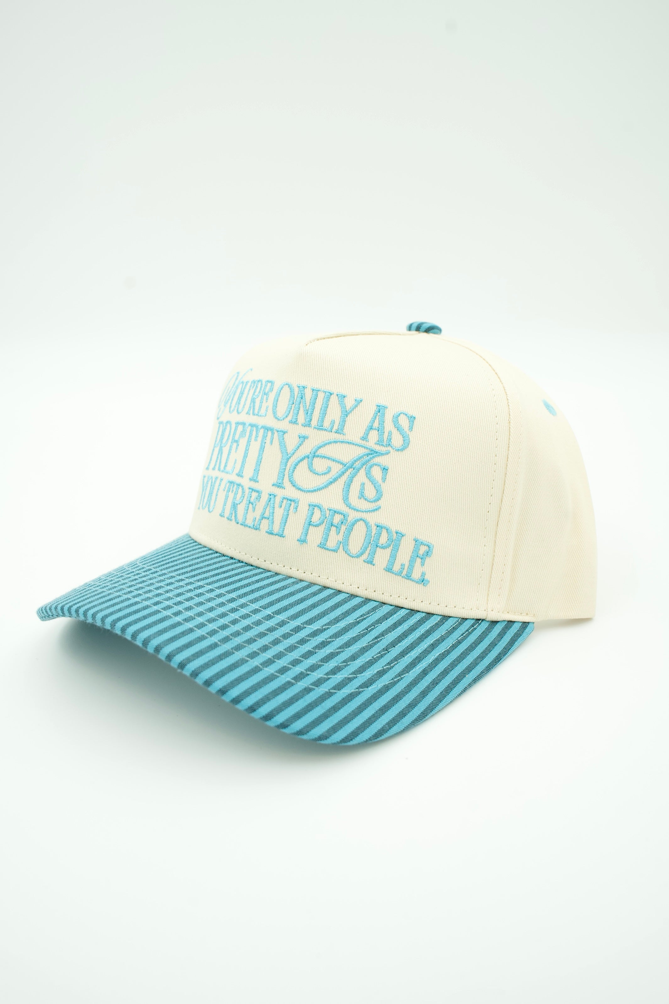 A cream-colored baseball cap with a light blue patterned brim and light blue embroidered text on the front, photographed on a clean white background.