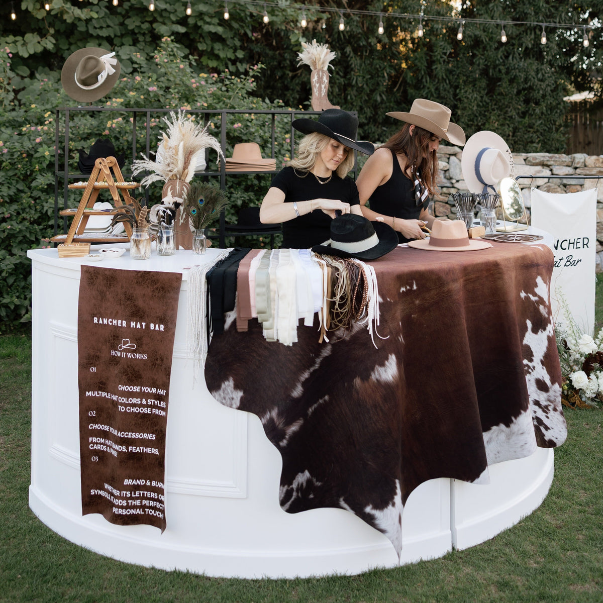 Outdoor event setup with two women seated behind a white circular booth draped in a brown-and-white cowhide blanket. The booth displays hats, woven textiles, and signage reading 'Handmade Hat Bar.