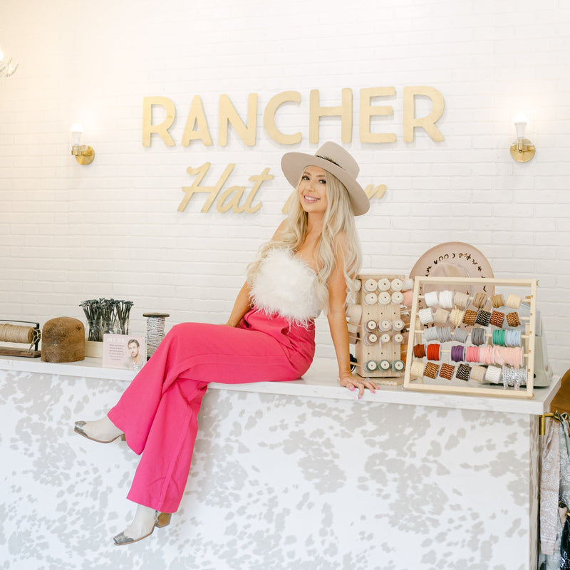 Shantelle Girard, CEO of Rancher Hat Bar sitting on a hat bar in a custom cowboy hat, a white feather shirt and pink dress pants