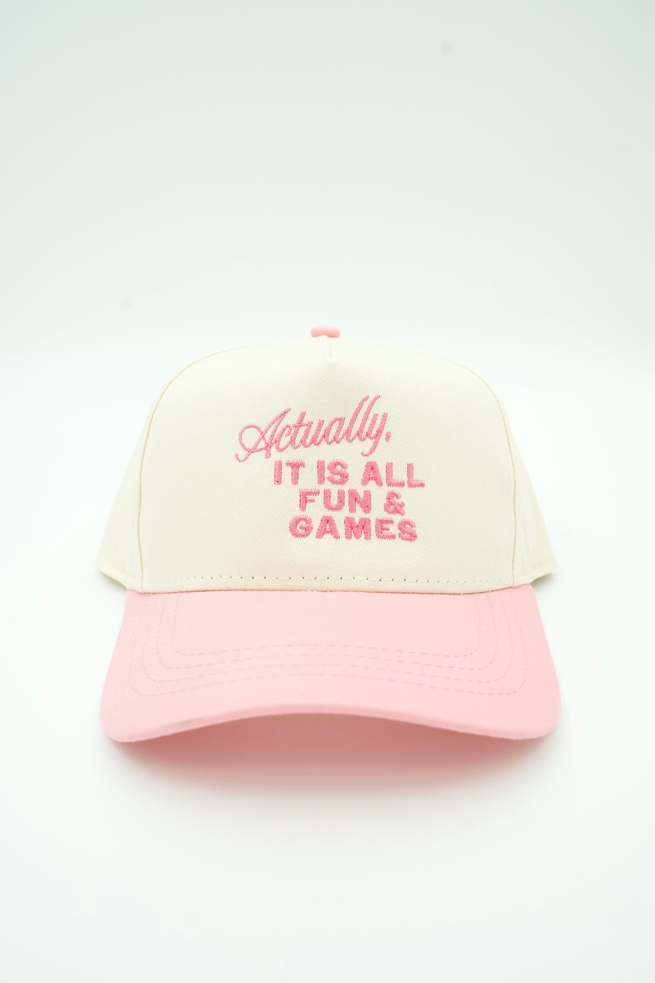A two-tone baseball cap with an off-white crown and a light pink brim, featuring pink embroidered text on the front that reads, 'Actually, IT IS ALL FUN & GAMES'.