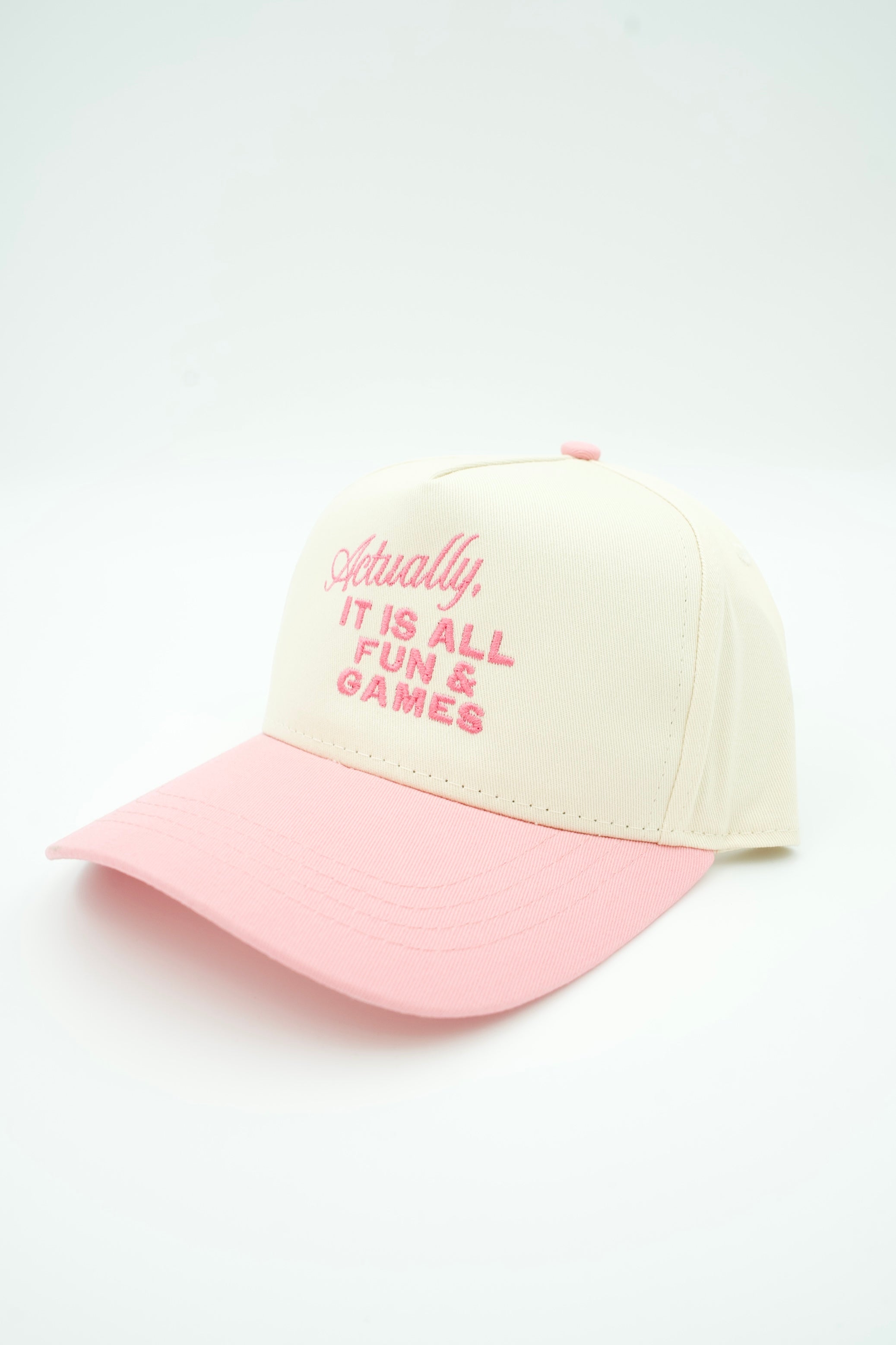 A baseball cap with a cream-colored crown and a light pink brim, featuring pink embroidered text on the front that reads 'Actually, IT IS ALL FUN & GAMES'.