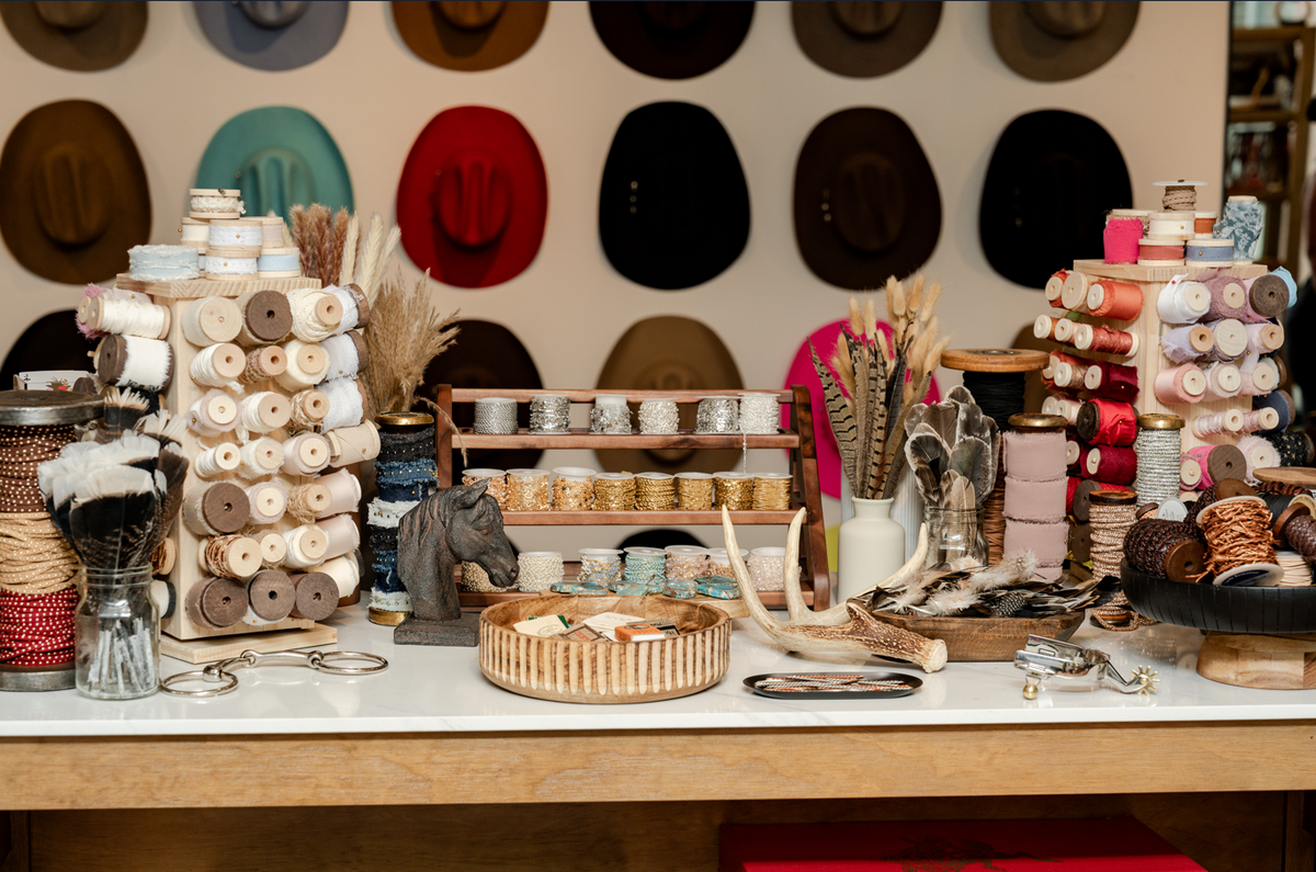 A styled display of craft supplies and textiles on a wooden table, featuring spools of thread, yarn, feathers, antlers, and small decorative items, with a wall of colorful hats in the background.
