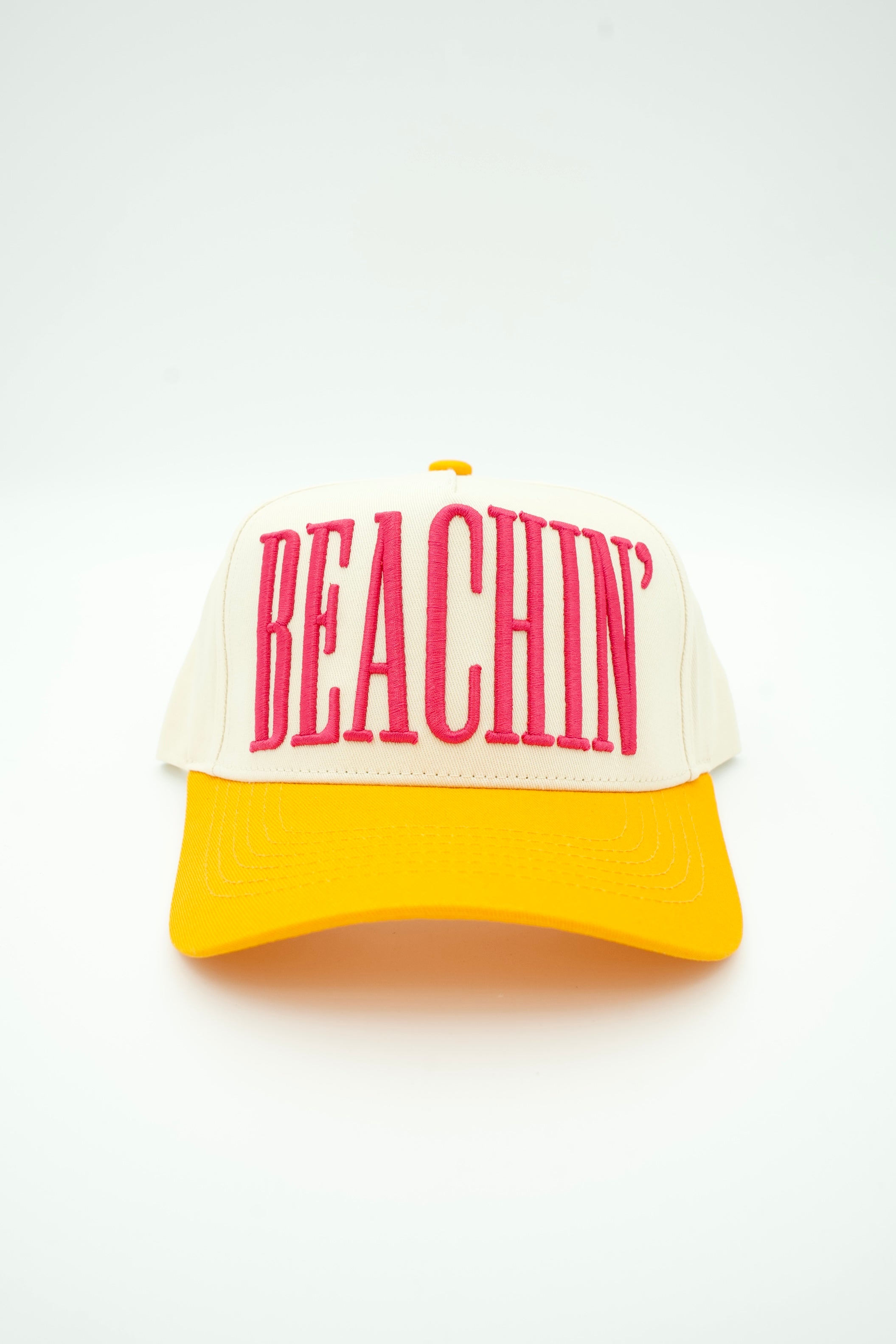 A two-tone trucker-style baseball cap with an off-white front panel and brim, featuring the word 'BEACHIN'' embroidered in bold pink letters across the front, and a bright yellow brim.
