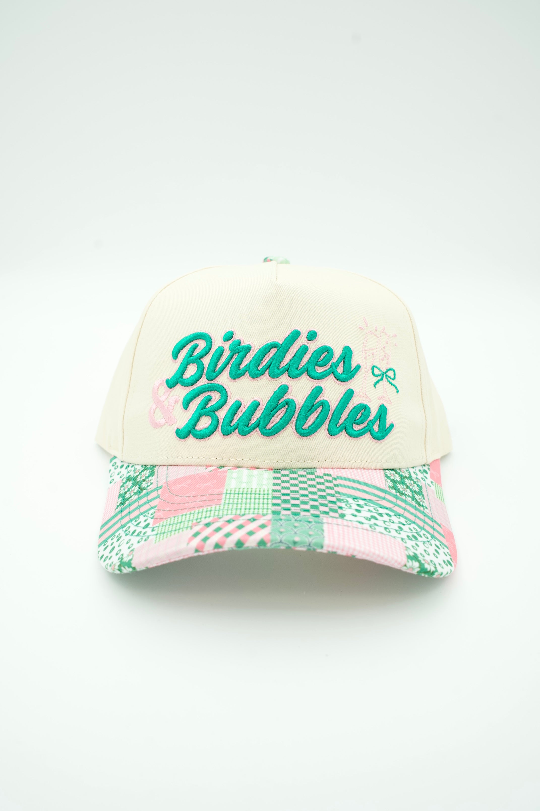 A baseball cap with a cream-colored front panel and a multicolor patchwork patterned brim. The front panel features the text 'Birdies Bubbles' in green script with a small bow graphic.