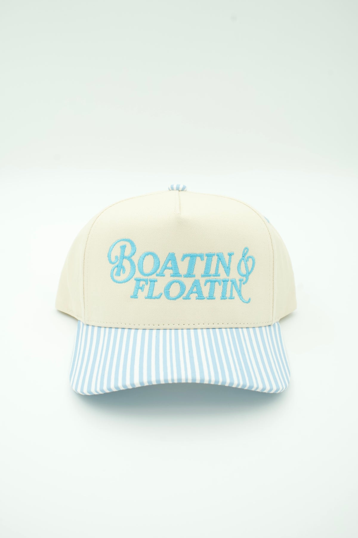 A cream-colored baseball cap with a light blue and white striped visor, featuring the words 'Boatin' & Floatin'' embroidered in light blue on the front.
