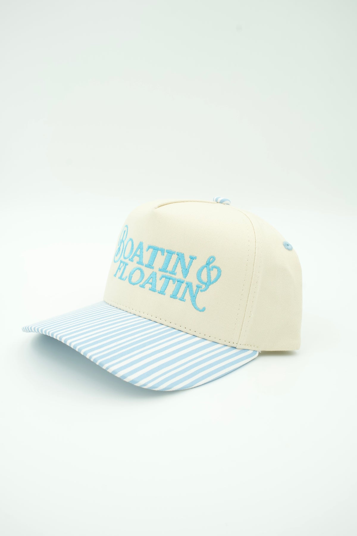 A baseball cap with a cream-colored crown, light blue embroidered text reading 'BOATIN' & FLOATIN'', and a light blue and white striped visor, shown on a plain white background.