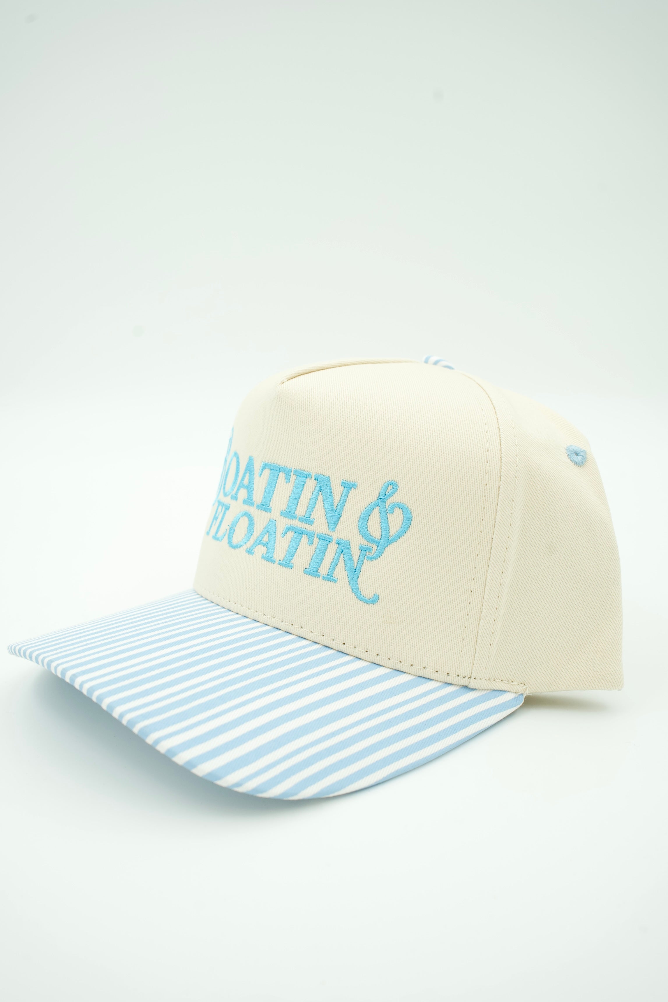 A baseball cap with an off-white crown, light blue embroidered text reading 'BOATIN' & FLOATIN'', and a light blue and white striped brim, shown on a plain light background.