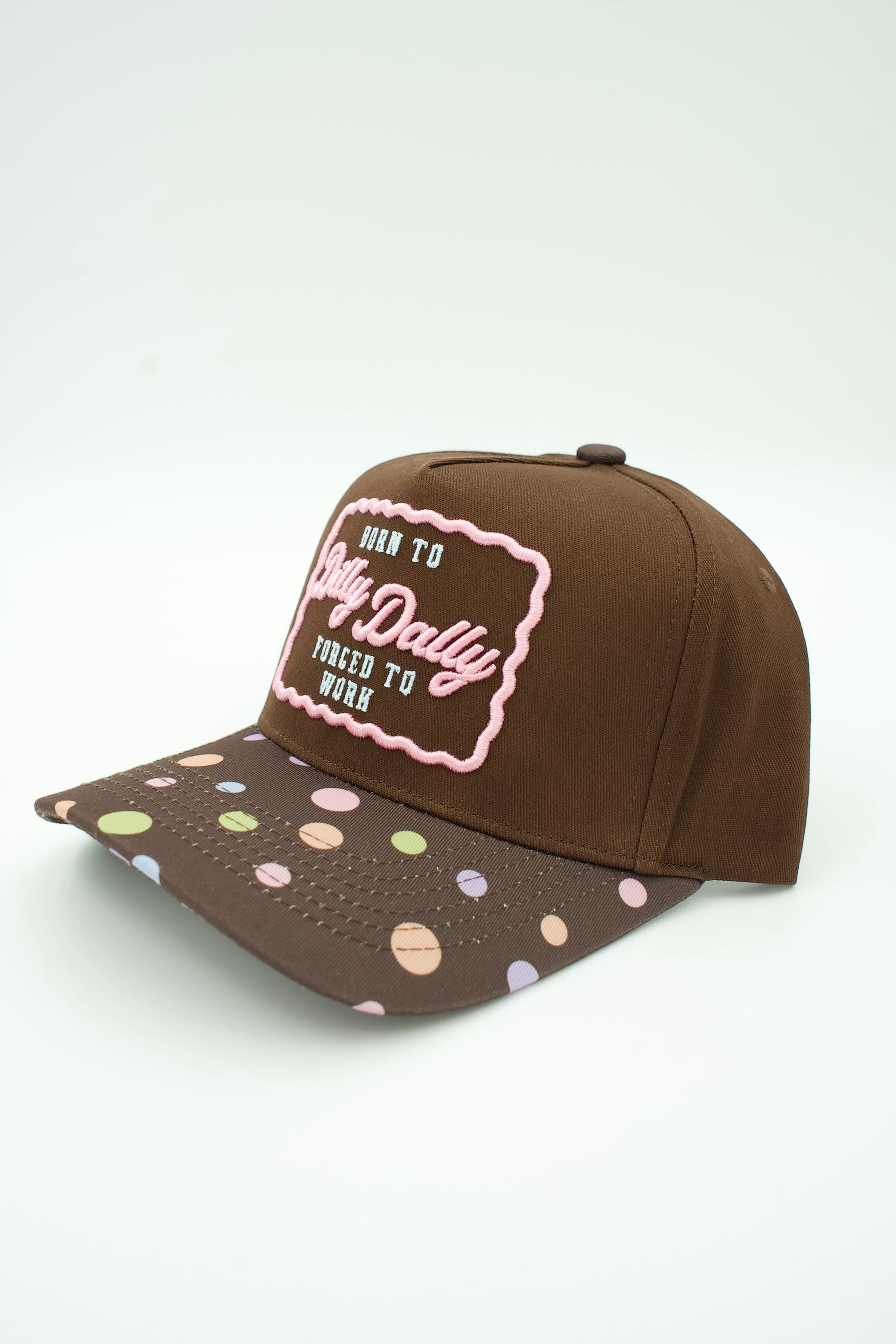 A brown baseball cap with a patterned visor featuring pastel-colored polka dots and a pink and blue embroidered patch on the front Born To Dilly Dally Forced To Work.