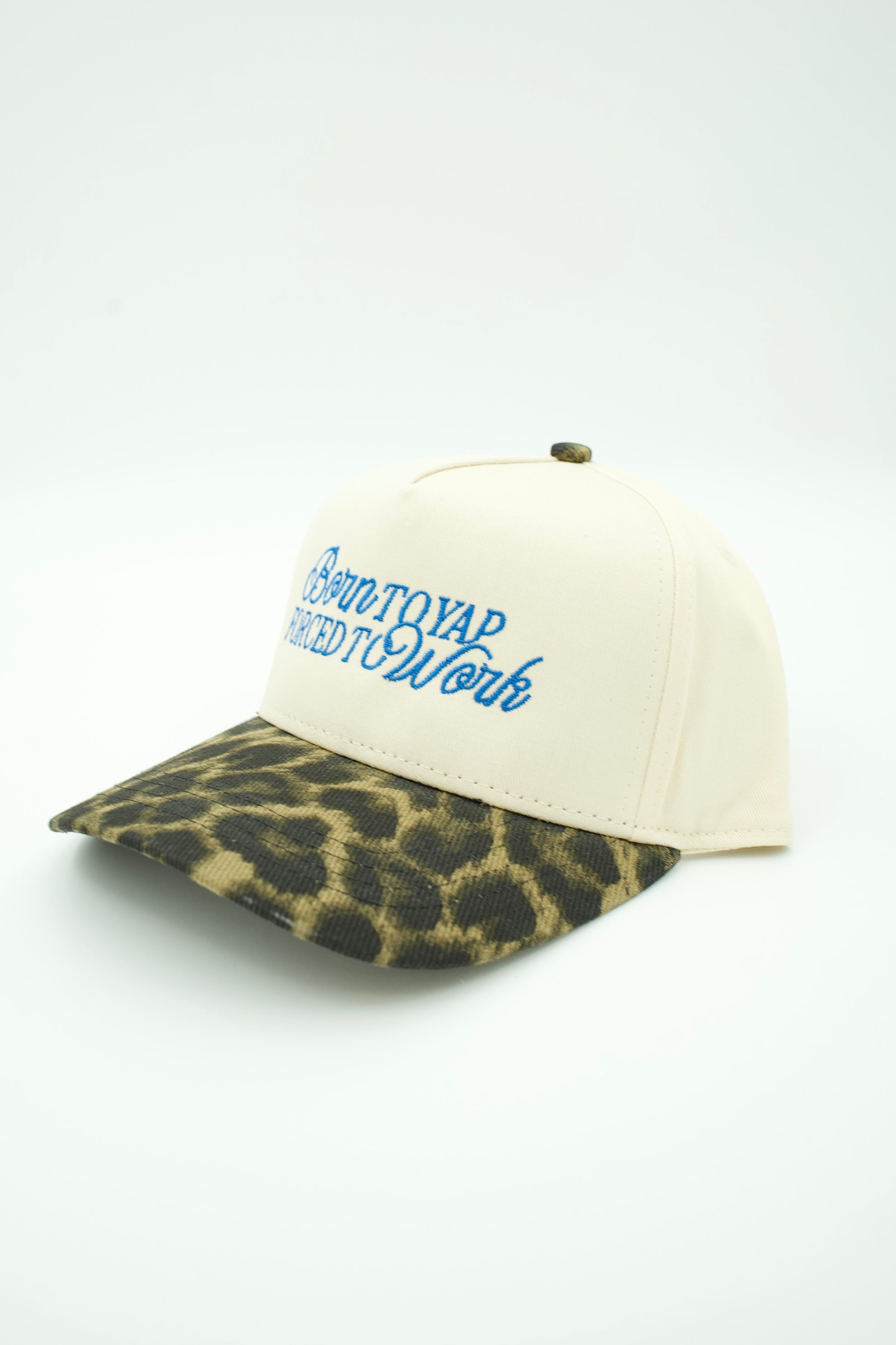 An off-white baseball cap with a leopard-print visor and blue embroidered text reading 'Born to Yap Forced to Work' on the front.