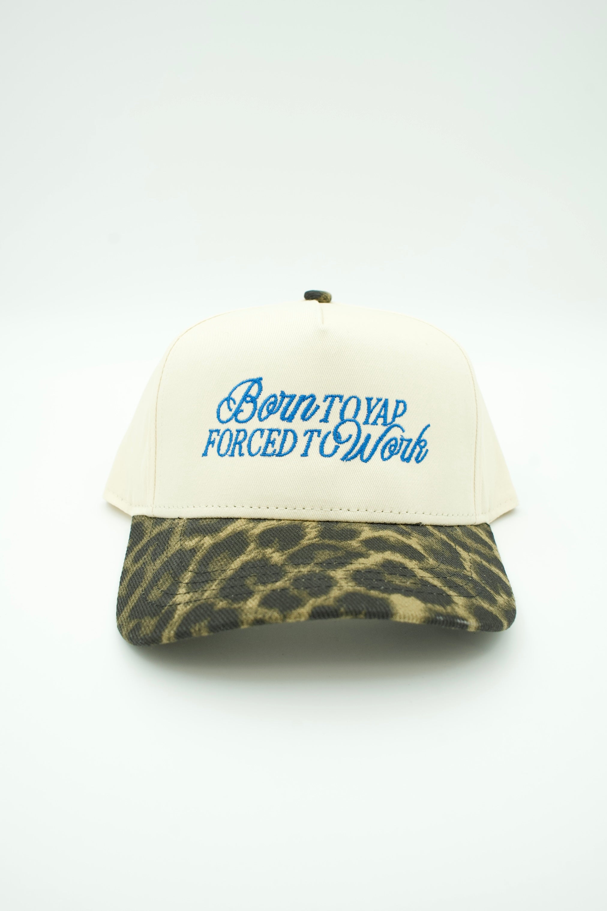 A baseball cap with an off-white crown and a leopard-print visor, featuring blue embroidered text on the front that reads 'Born to Yap Forced to Work'.