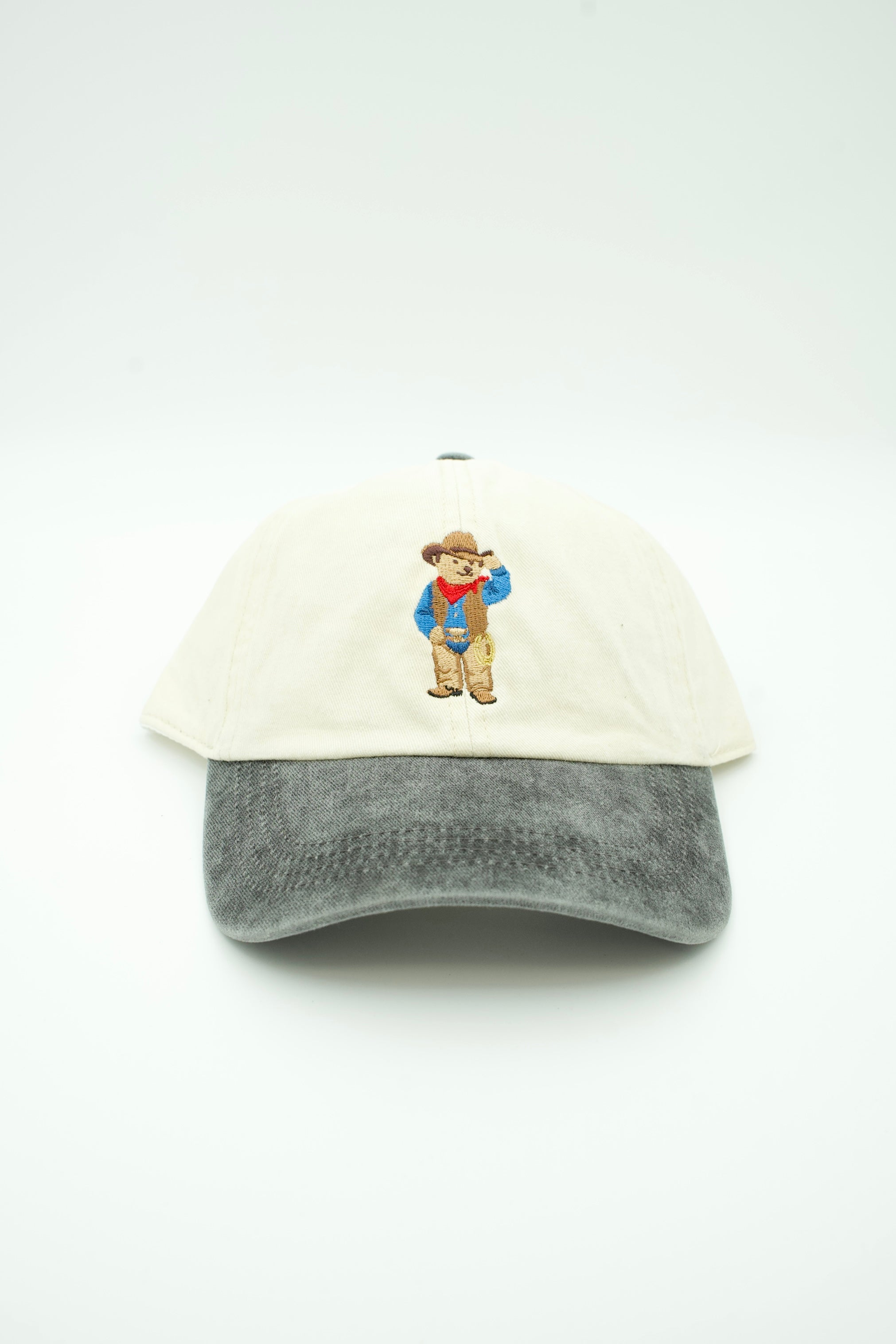 A trucker hat with an off-white crown and a gray washed brim, featuring a small embroidered cartoon bear wearing a cowboy hat and blue jacket on the front.